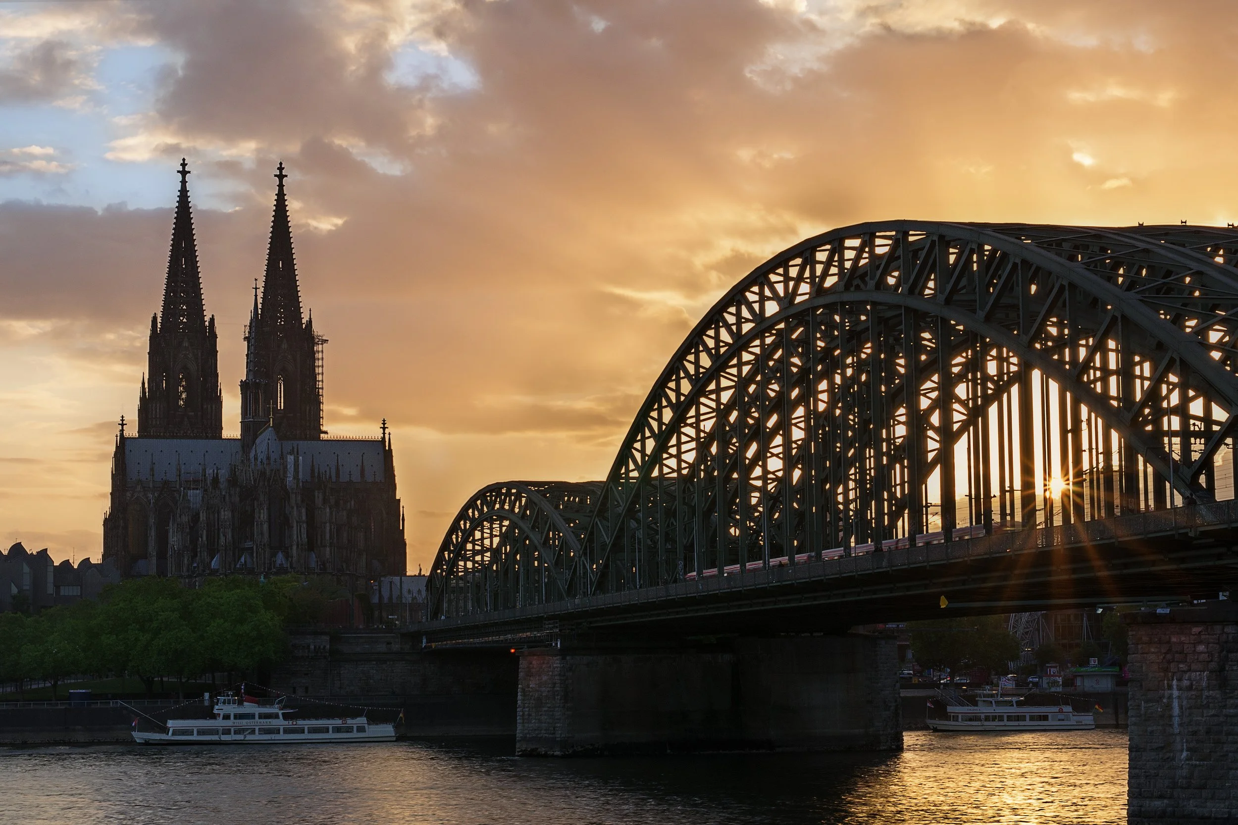 Sunset over the Cologne Cathedral and Hohenzollern Bridge in Cologne, Germany, with a river in the foreground and boats on the water.