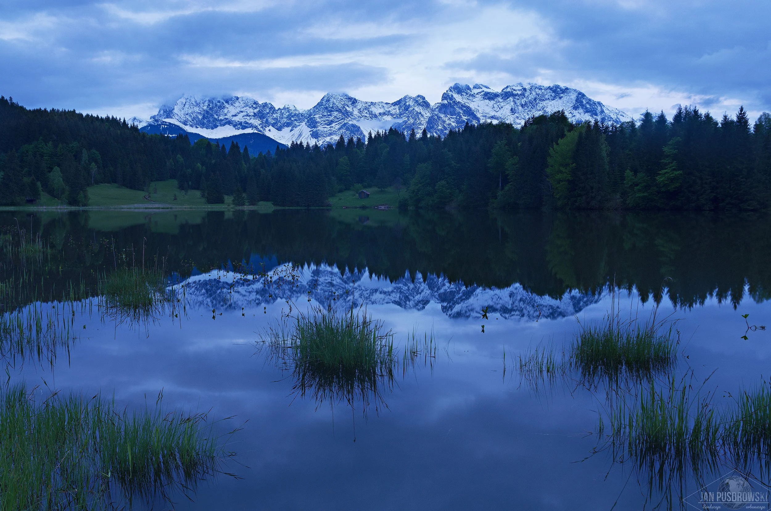Snow-capped mountains reflecting in a calm lake with dense forest and a cloudy sky. Barmsee Bayern, Bavaria, Germany, Deutschland.