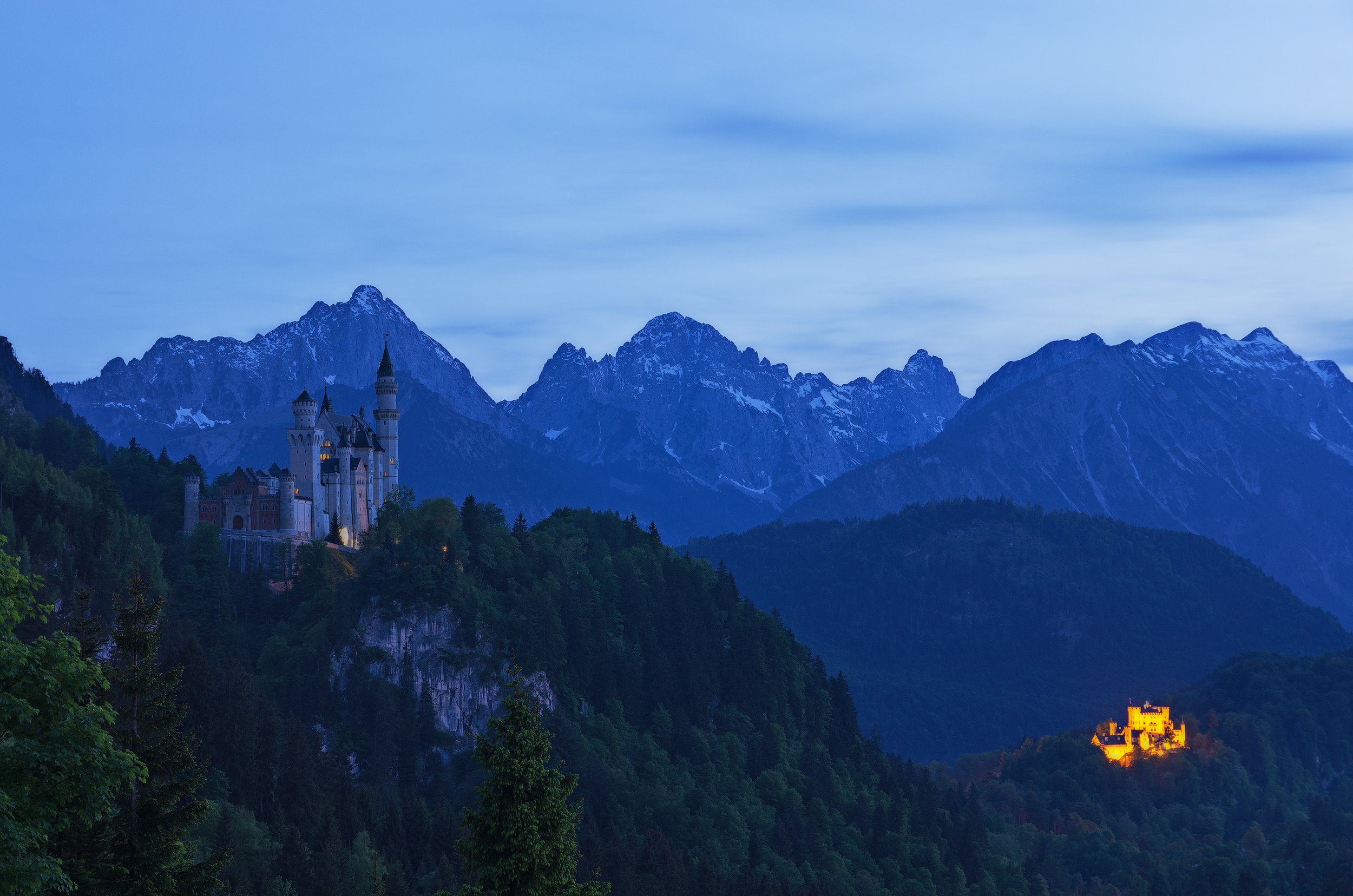 Nighttime view of a mountain landscape with a castle on a hill and another illuminated castle in the distance, surrounded by forested mountains.