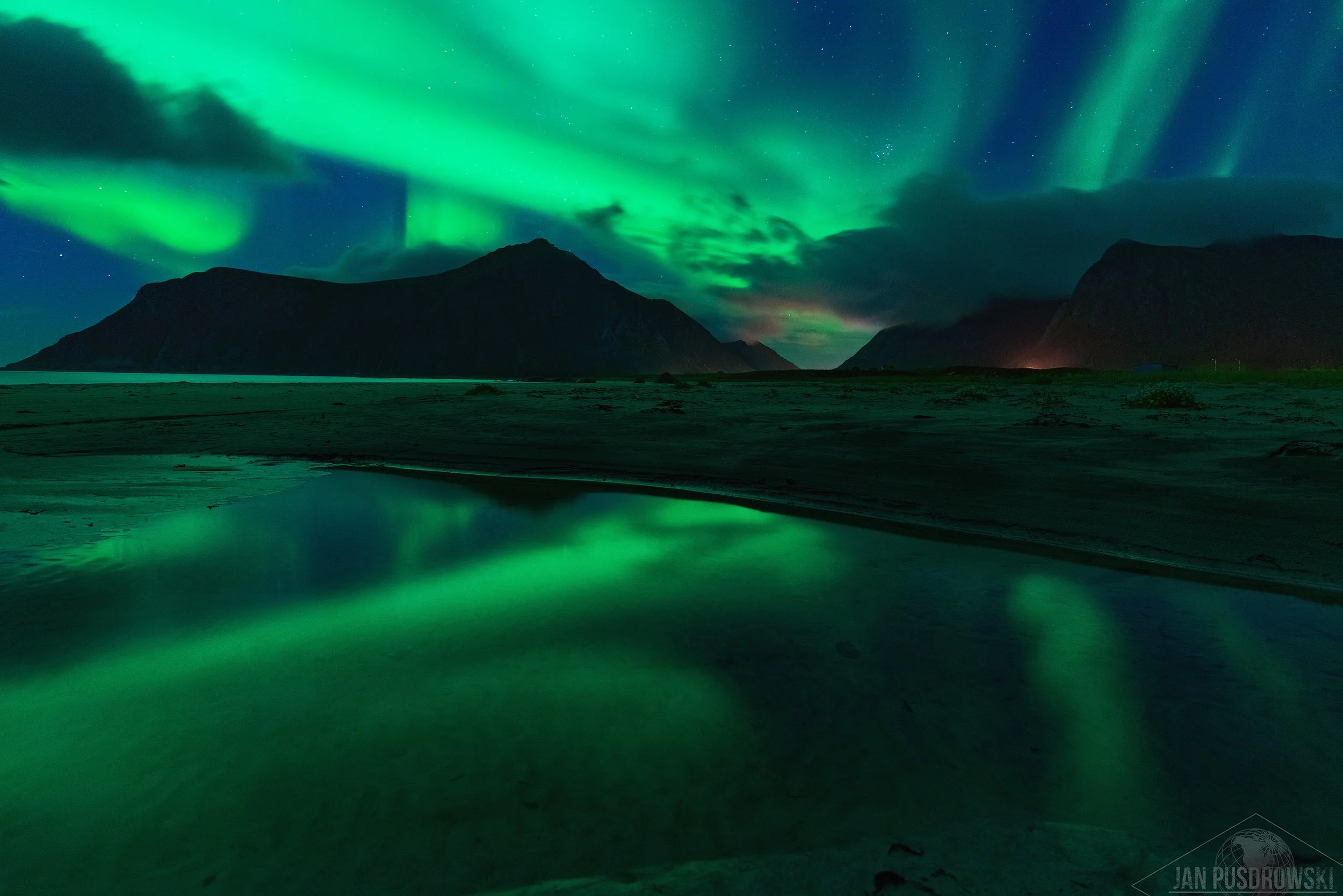 Northern lights illuminating the night sky over a mountain range and reflecting on a body of water. Lofoten Norway.