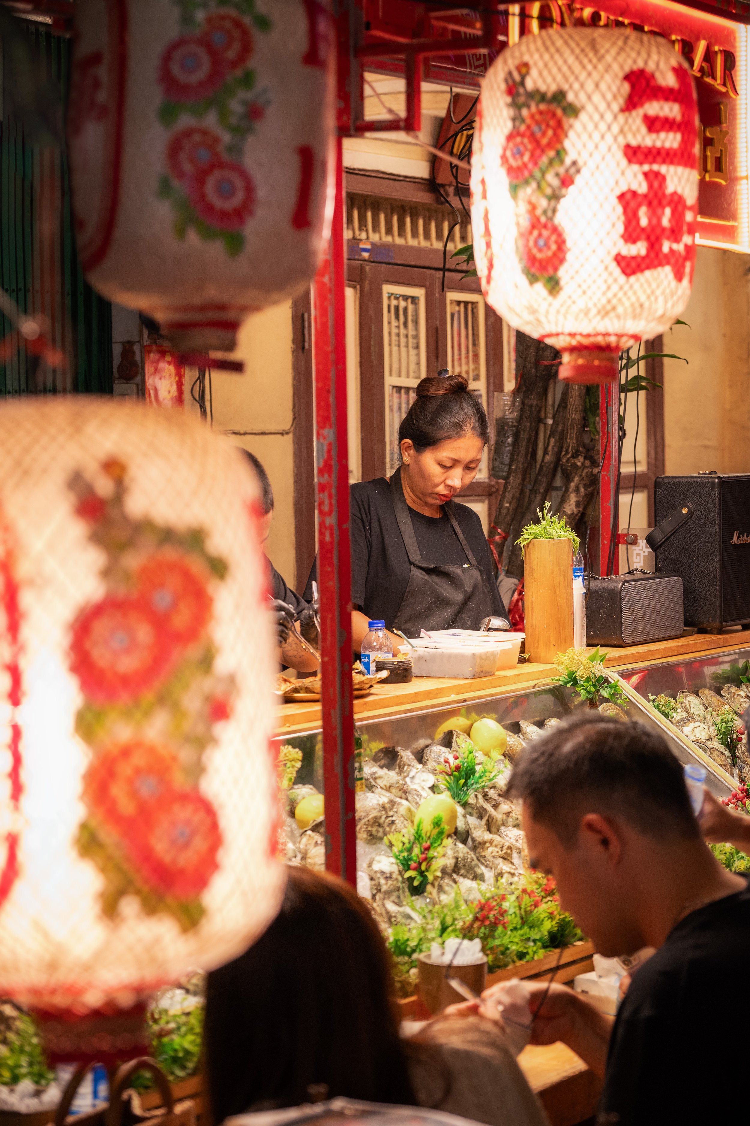 A woman stall owner at a food stand decorated with Chinese lanterns, selling seafood and vegetables in an outdoor market at night.