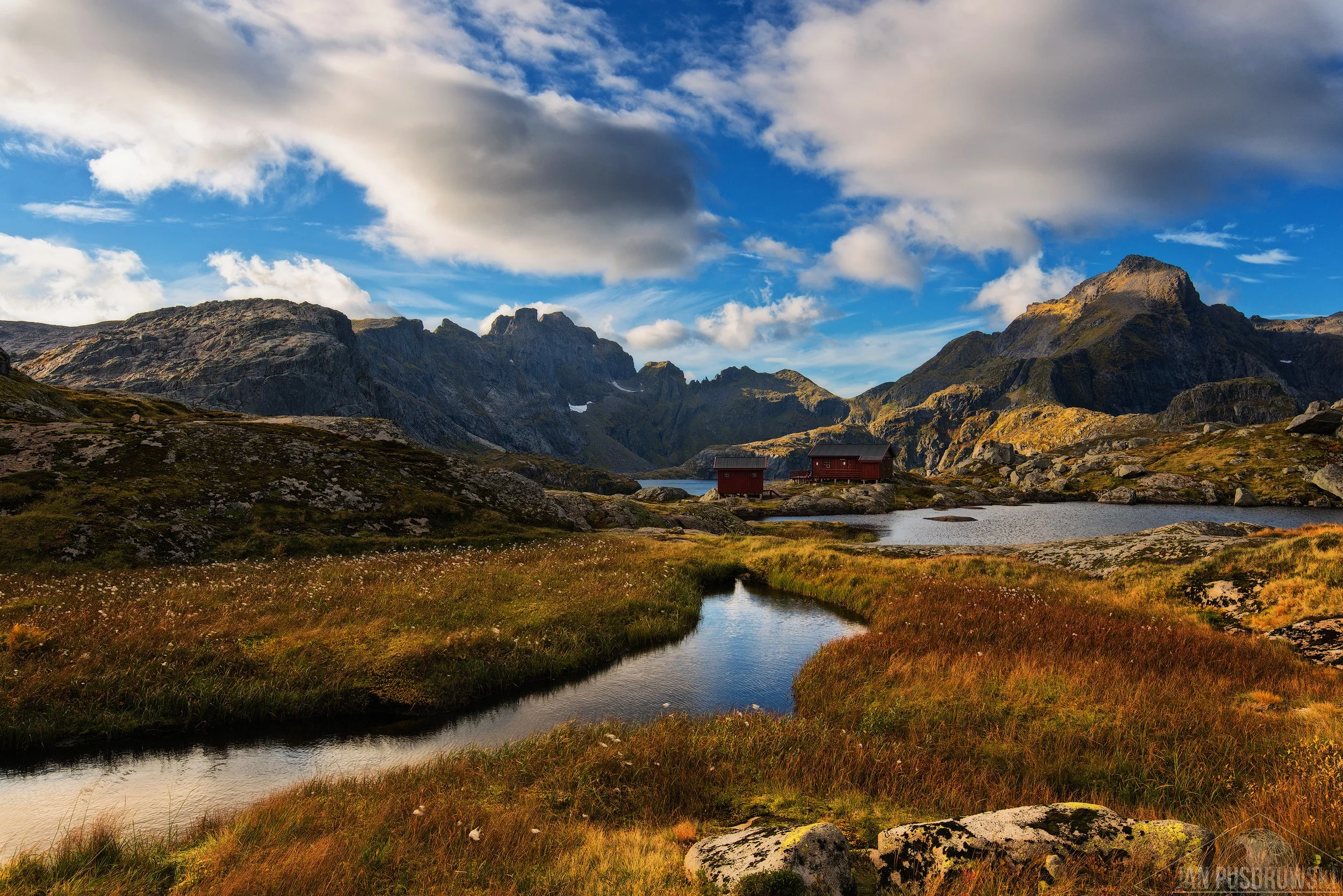 Scenic mountain landscape with a small river or stream in the foreground, grassy terrain, and two red cabins near a lake at the base of rugged mountains under a partly cloudy sky. Lofoten, Norway.