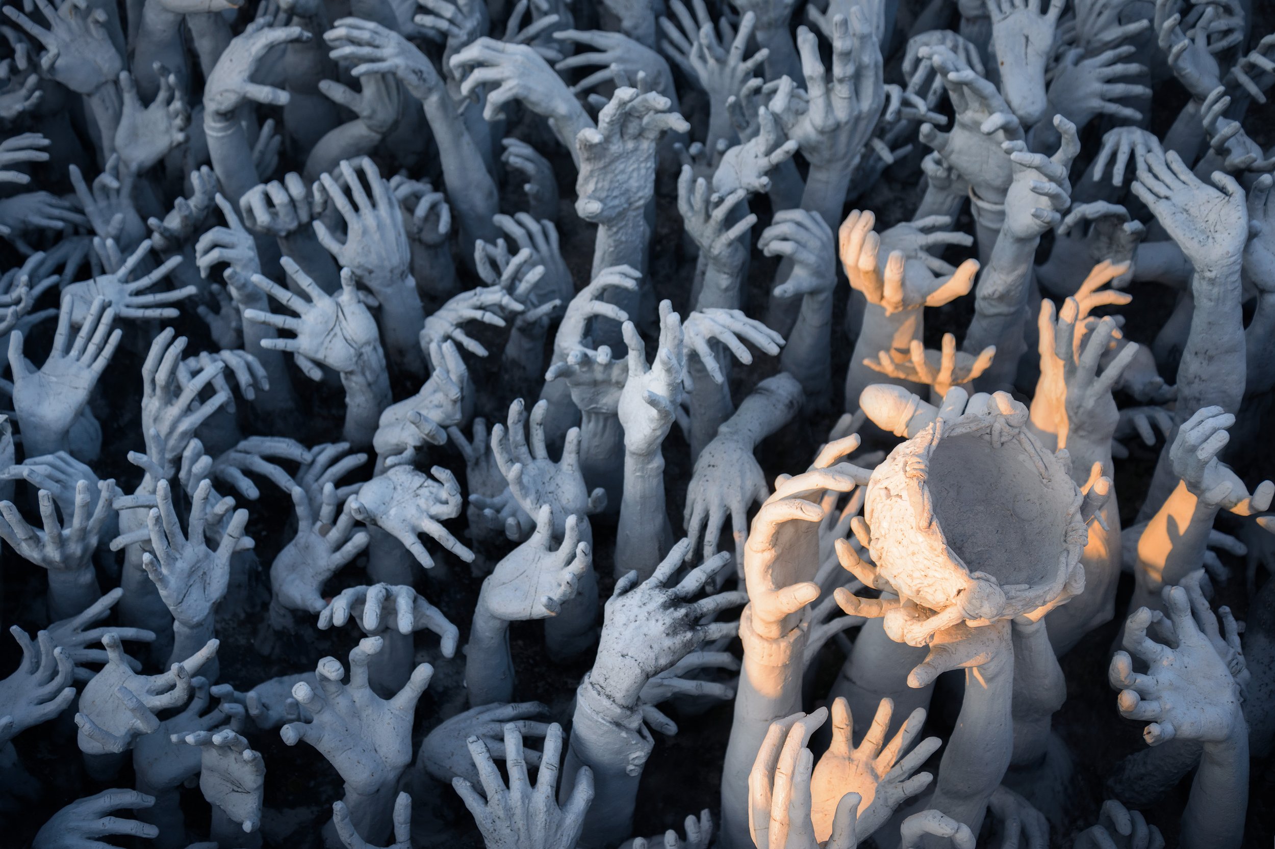 A large group of white, clay or plaster hands reaching up, with some holding a broken bowl, all emerging from the ground. Thailand Chiang Rai.
