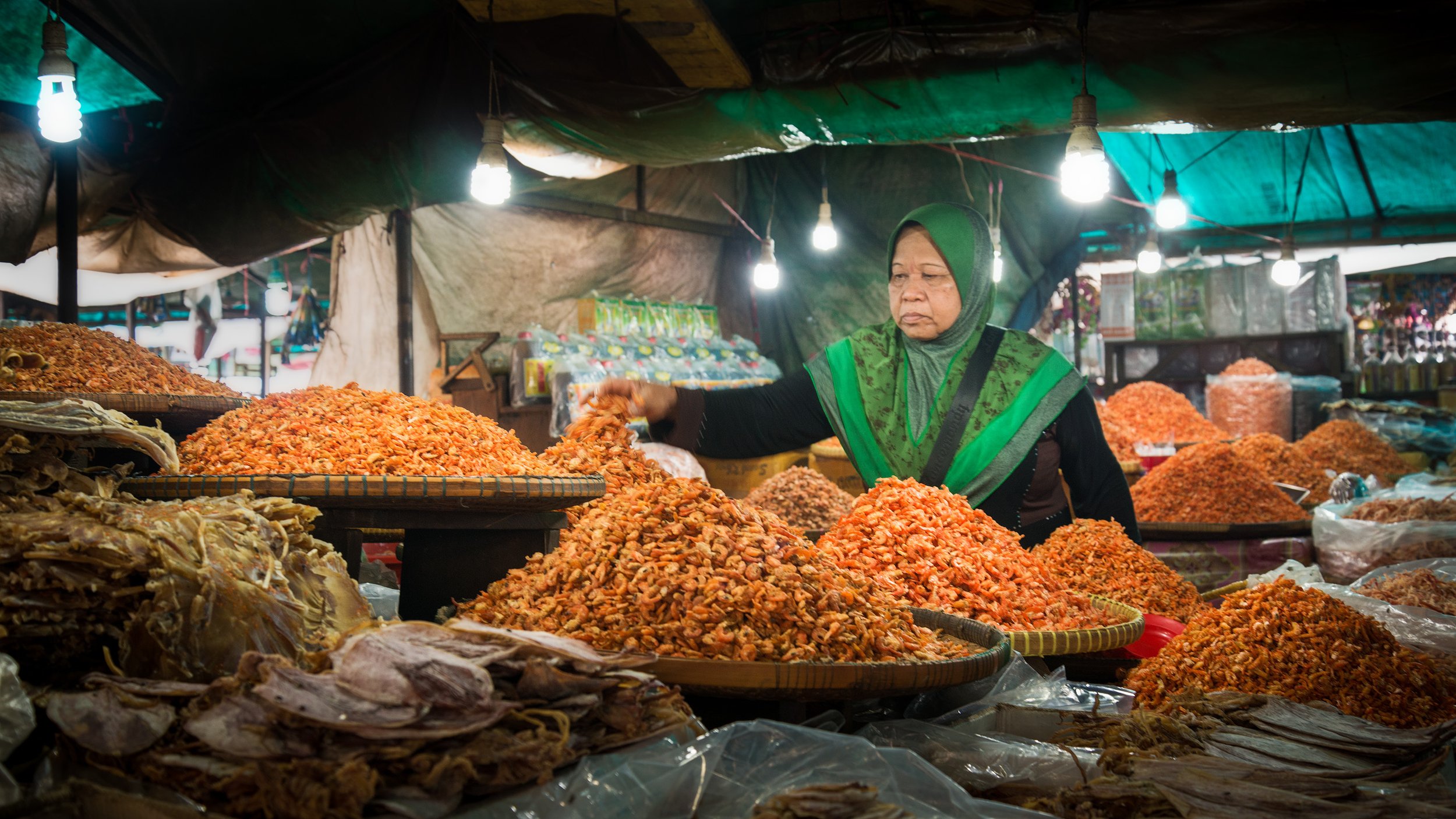 A woman wearing a green headscarf and black clothing sells various types of dried seafood at an outdoor market, with piles of dried shrimp and fish on display under hanging lights.