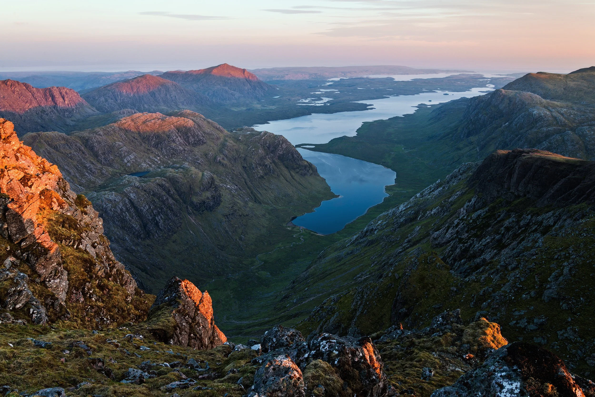 A view of a mountain valley with multiple lakes, rugged hills, and distant mountains under a pastel-colored sky at sunset.