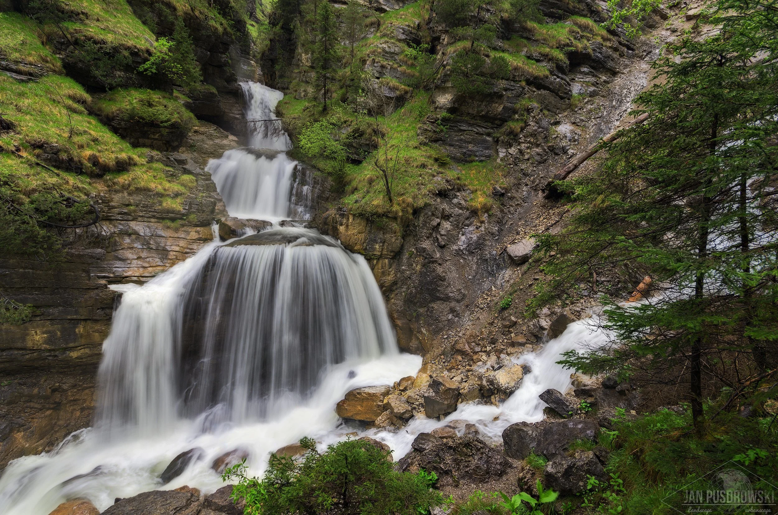 A waterfall cascading down rocky cliffs surrounded by lush green trees and vegetation. Kuhflucht, Bayern, Deutschland.