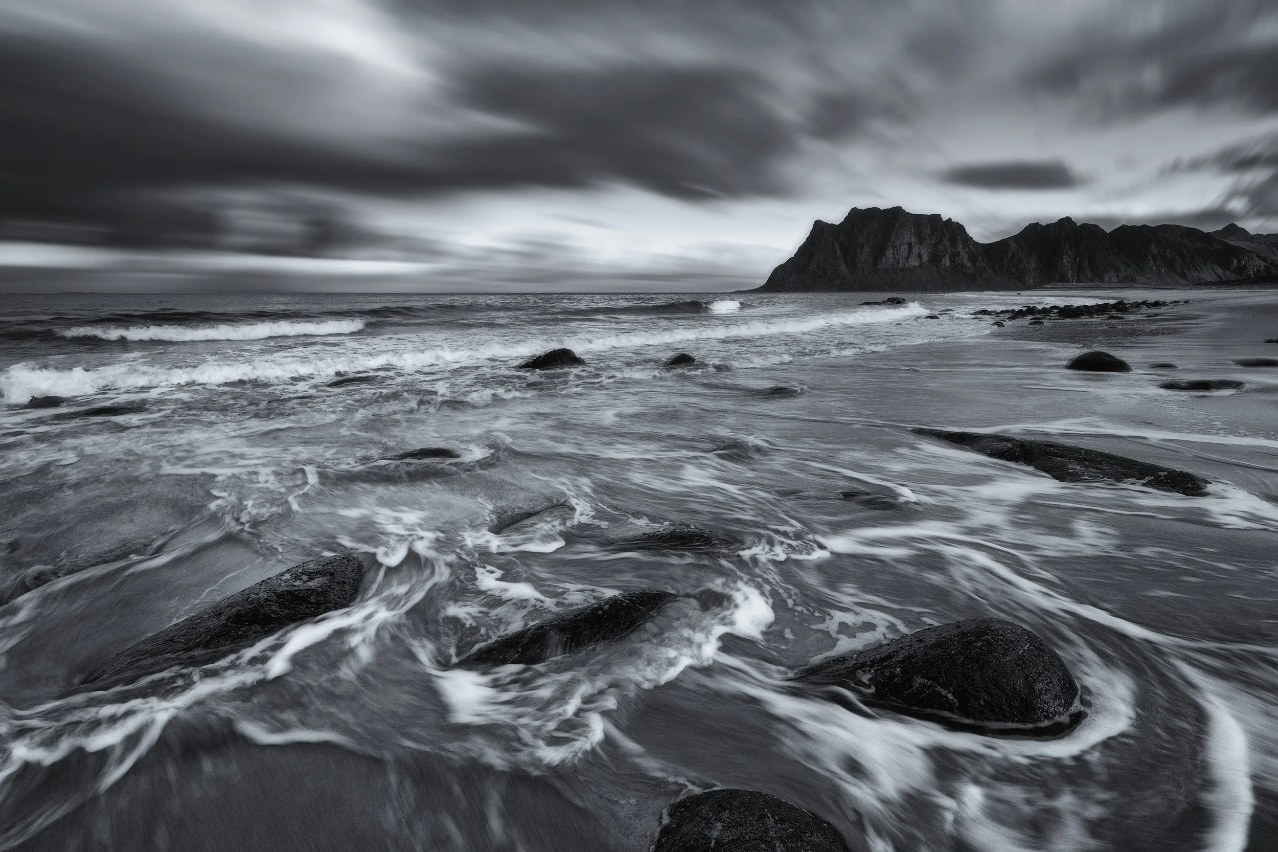 Black and white photo of a rocky beach with waves crashing and a mountain in the background under a cloudy sky. Lofoten Norway.