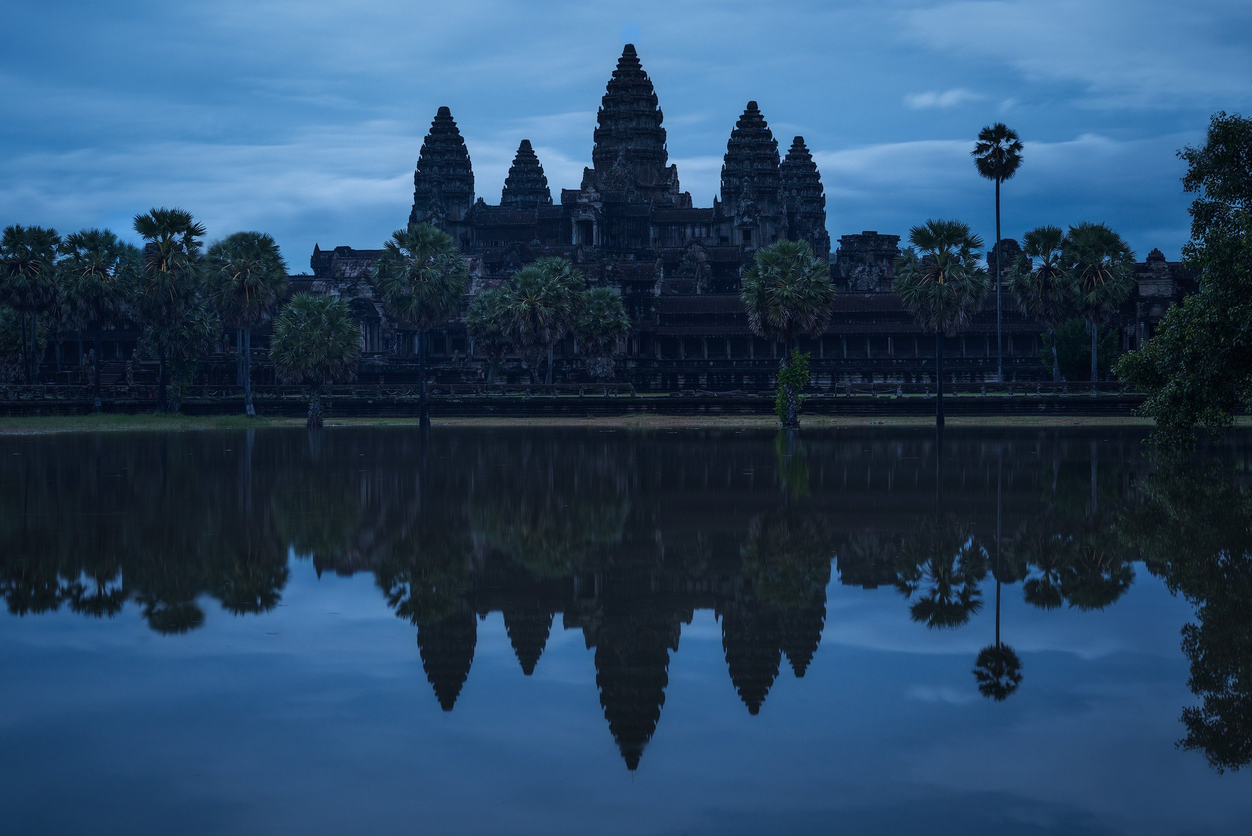 Silhouette of Angkor Wat temple at dawn reflected in a body of water, surrounded by palm trees and with a cloudy sky in the background. Angkor wat Cambodia.