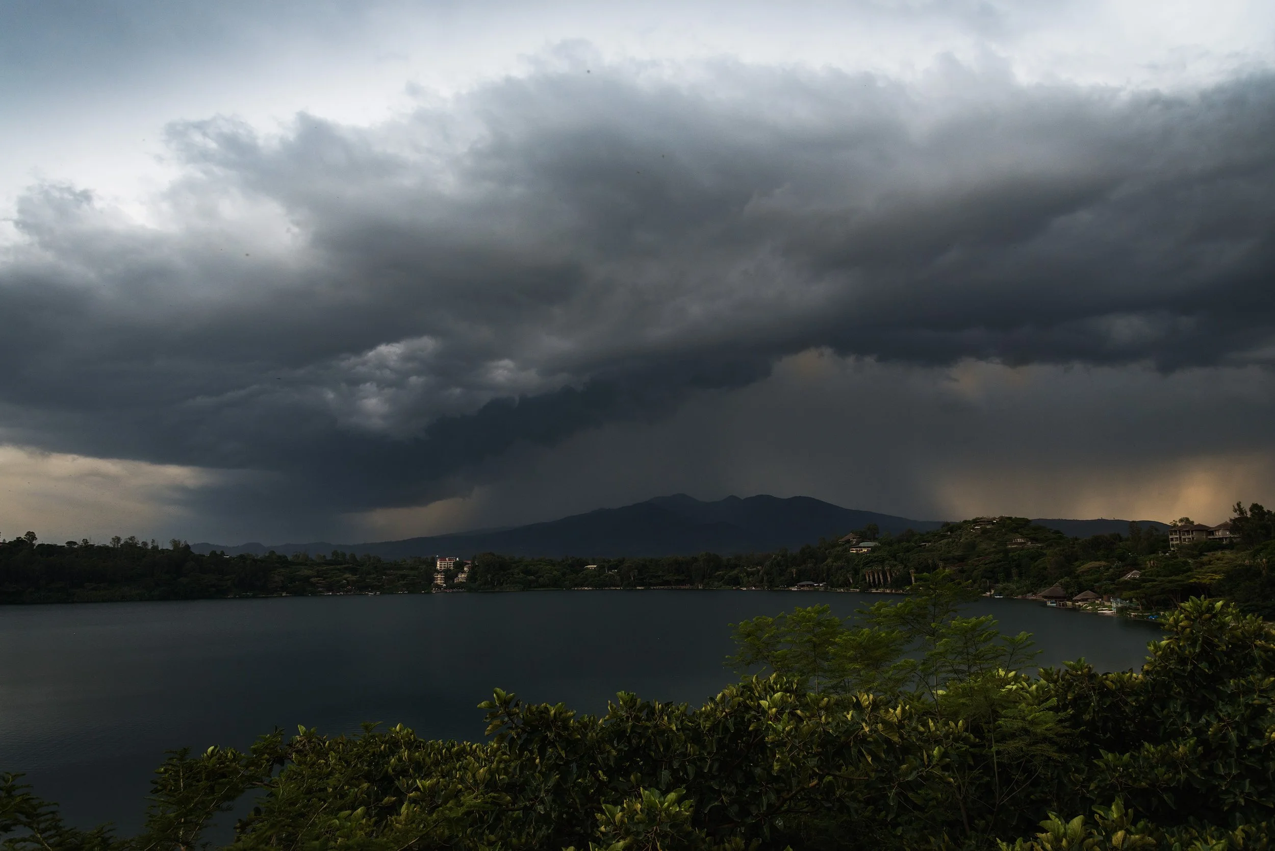 Dark storm clouds over a mountain range with rain falling in the distance, overlooking a large lake with green foliage in the foreground.