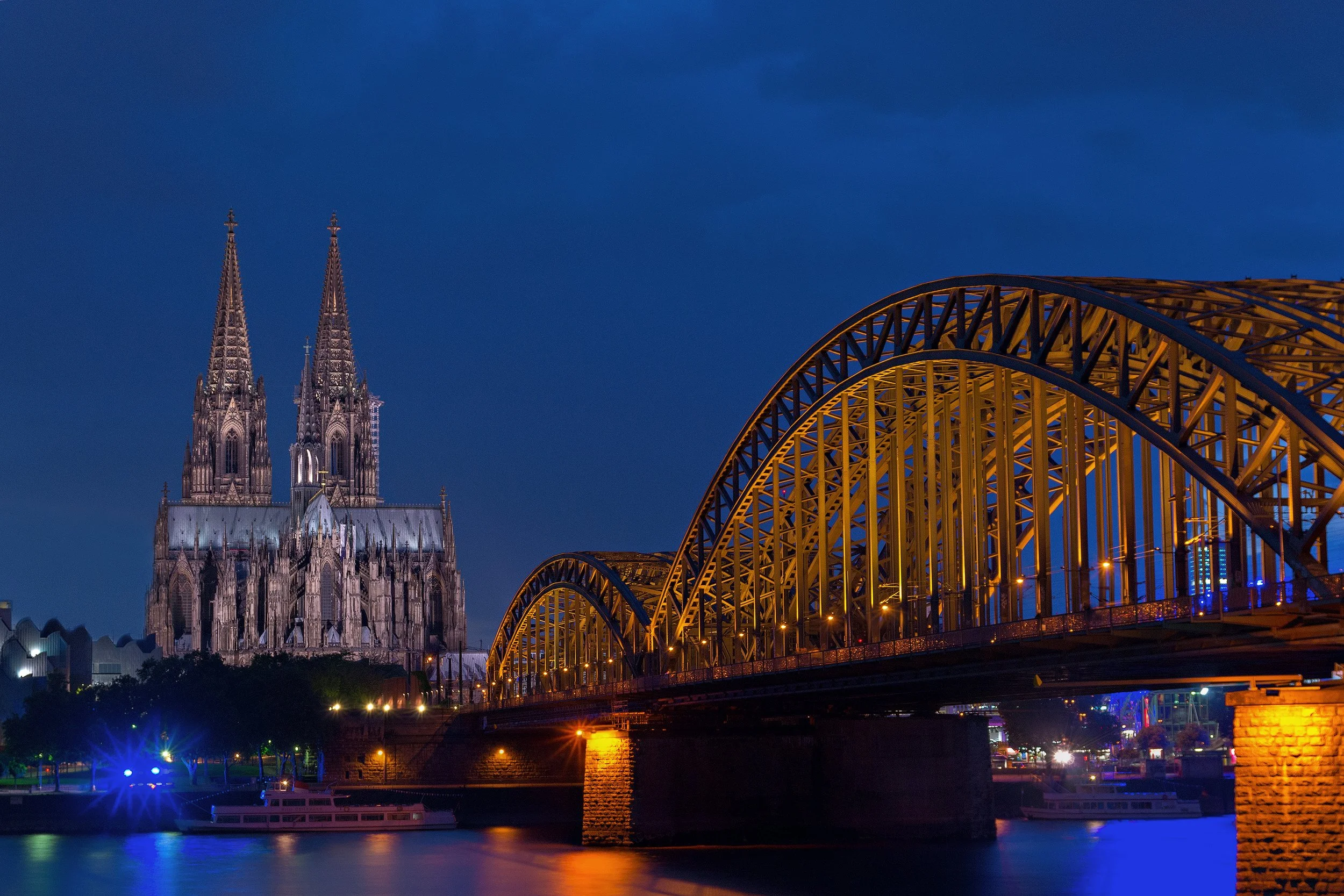 Nighttime view of a large Gothic cathedral with twin spires, illuminated, standing next to an arched bridge with yellow lighting, over a river with boats, and a dark blue sky.