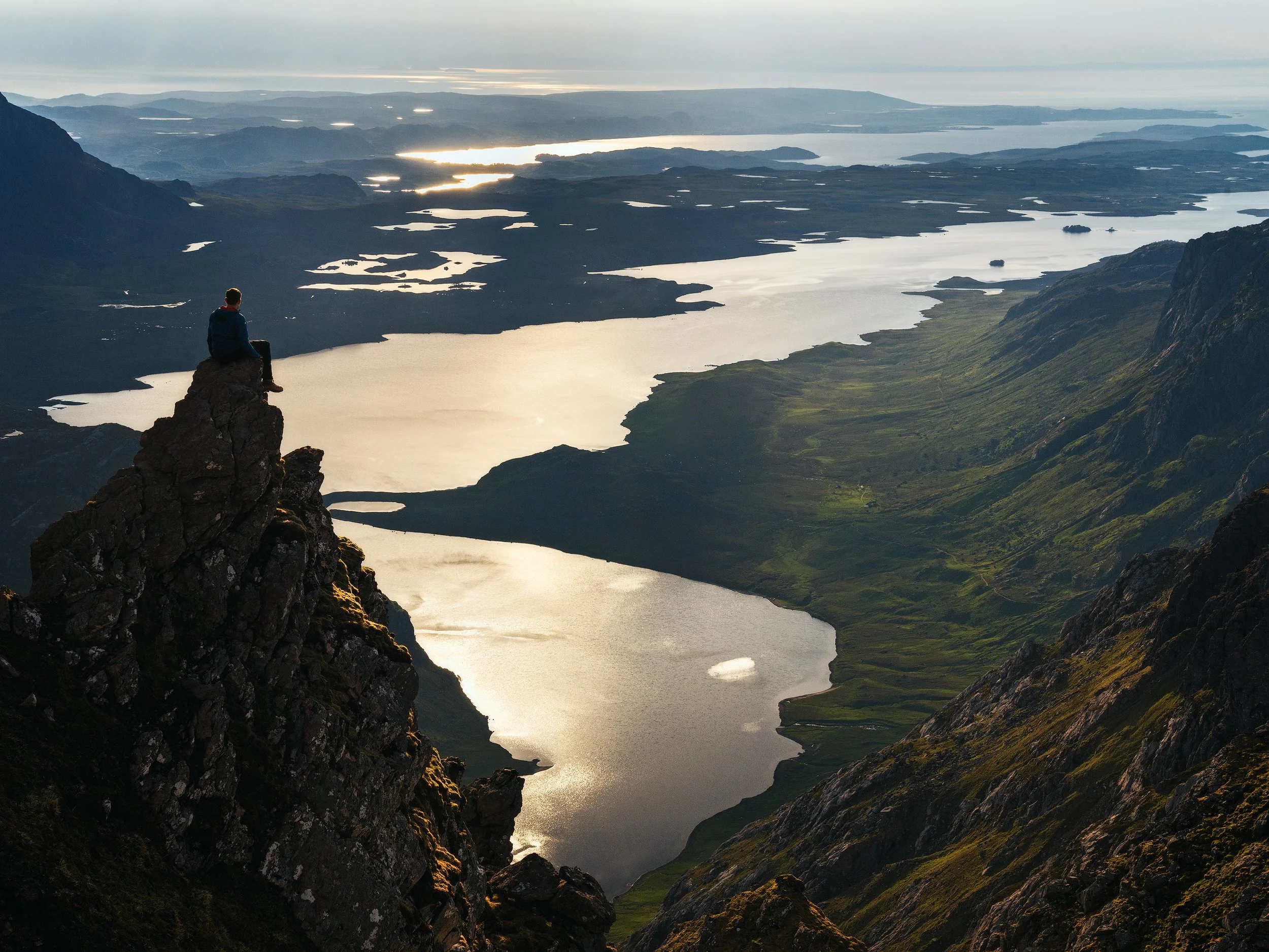 A person sitting on a rocky cliff overlooking a large lake and mountain landscape during sunset.