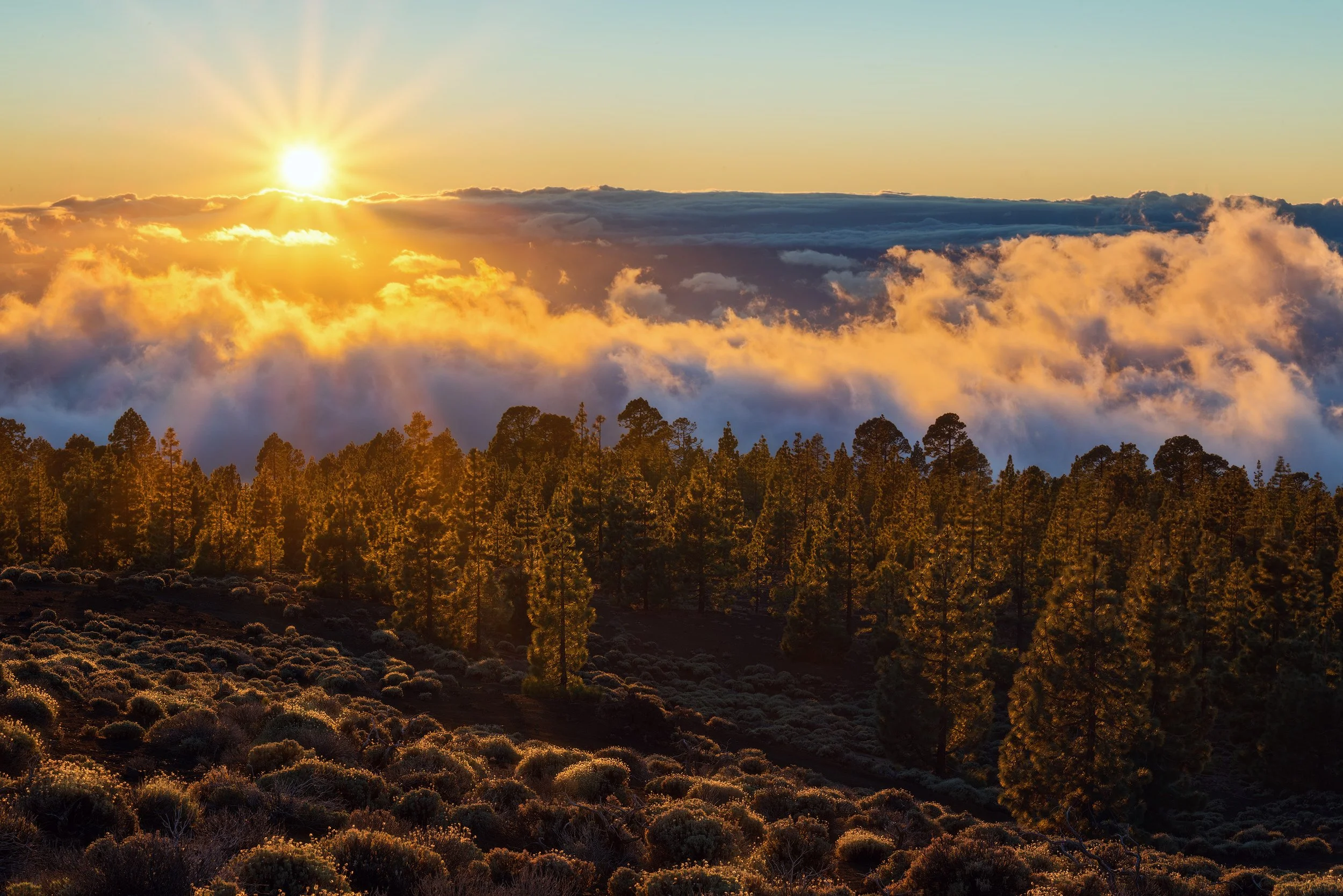 Sunset over a forested mountain landscape with clouds and rays of sunlight.