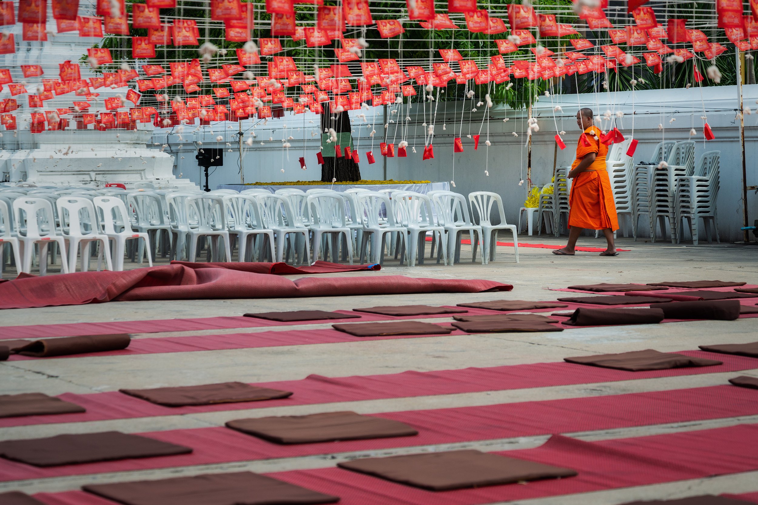 Buddhist monk in orange robes walking past rows of chairs and mats, with red banners hanging overhead, set up for a ceremony on an outdoor platform.