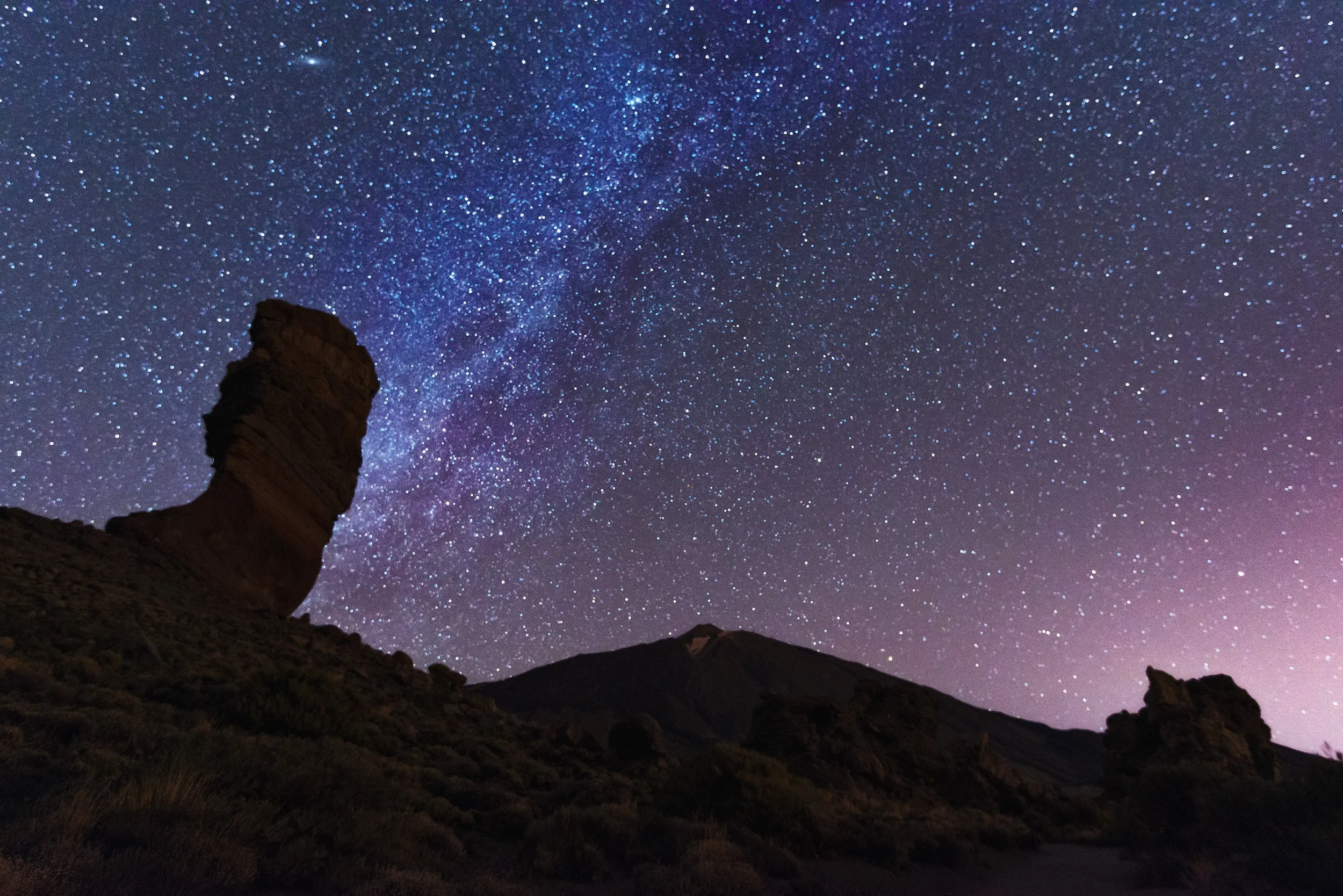 Night sky filled with stars and the Milky Way galaxy over a rugged landscape with rock formations.