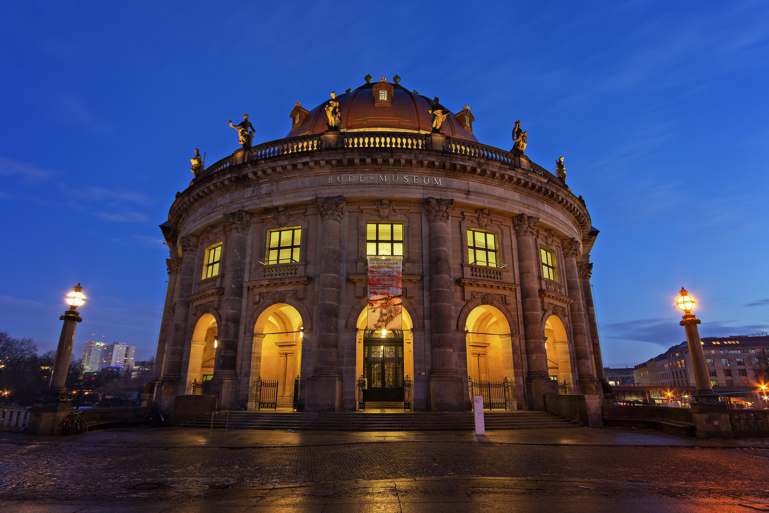 The Bode Museum in Berlin illuminated at dusk, showcasing its neoclassical architecture with large arched entrances, columns, and decorative statues on the roof, against a darkening blue sky.