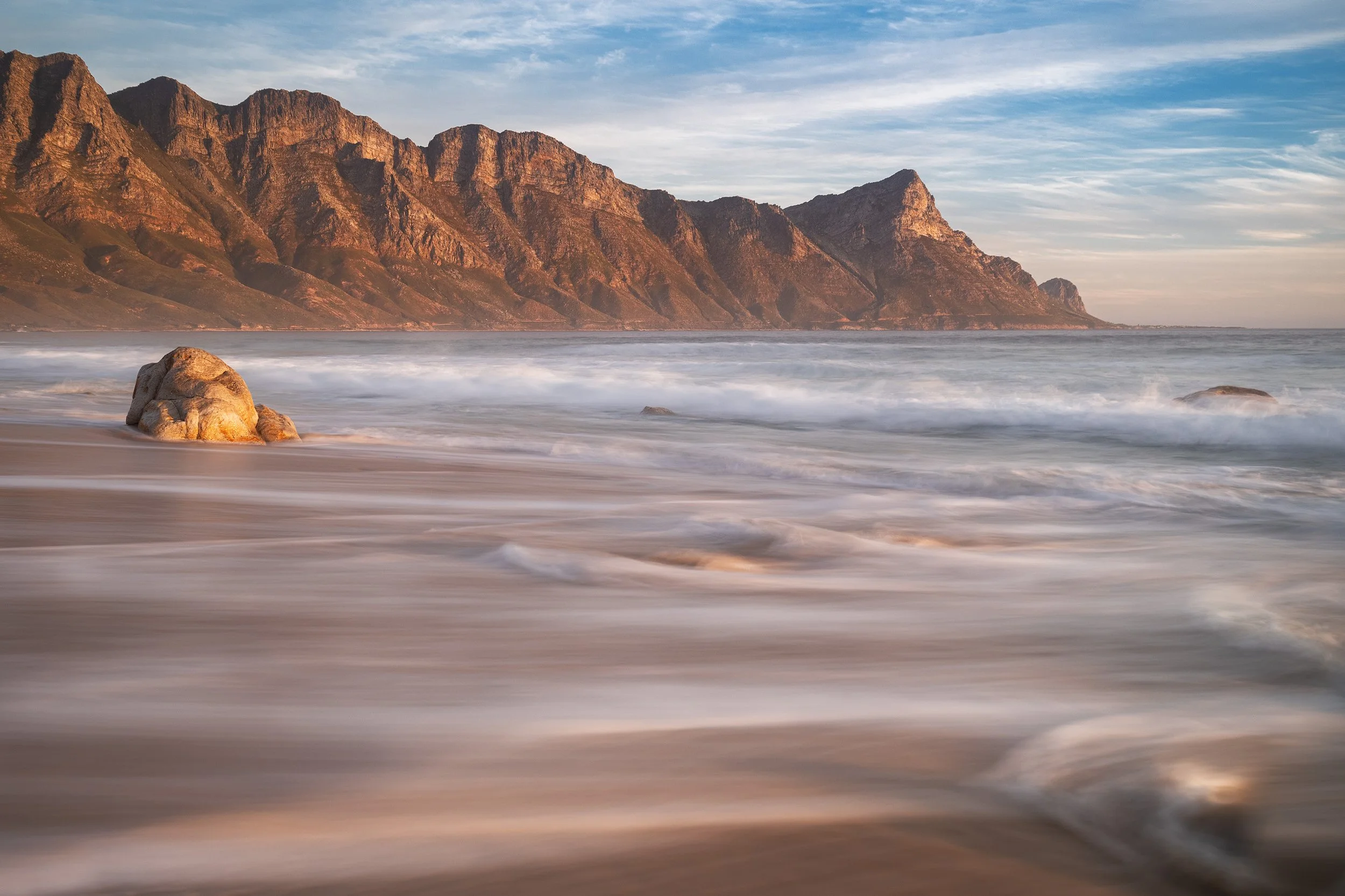 A scenic beach with a large rock in the foreground, shoreline waves, and rugged mountains in the background under a sky with light clouds. Kogel bay, South Africa, Cape Town.