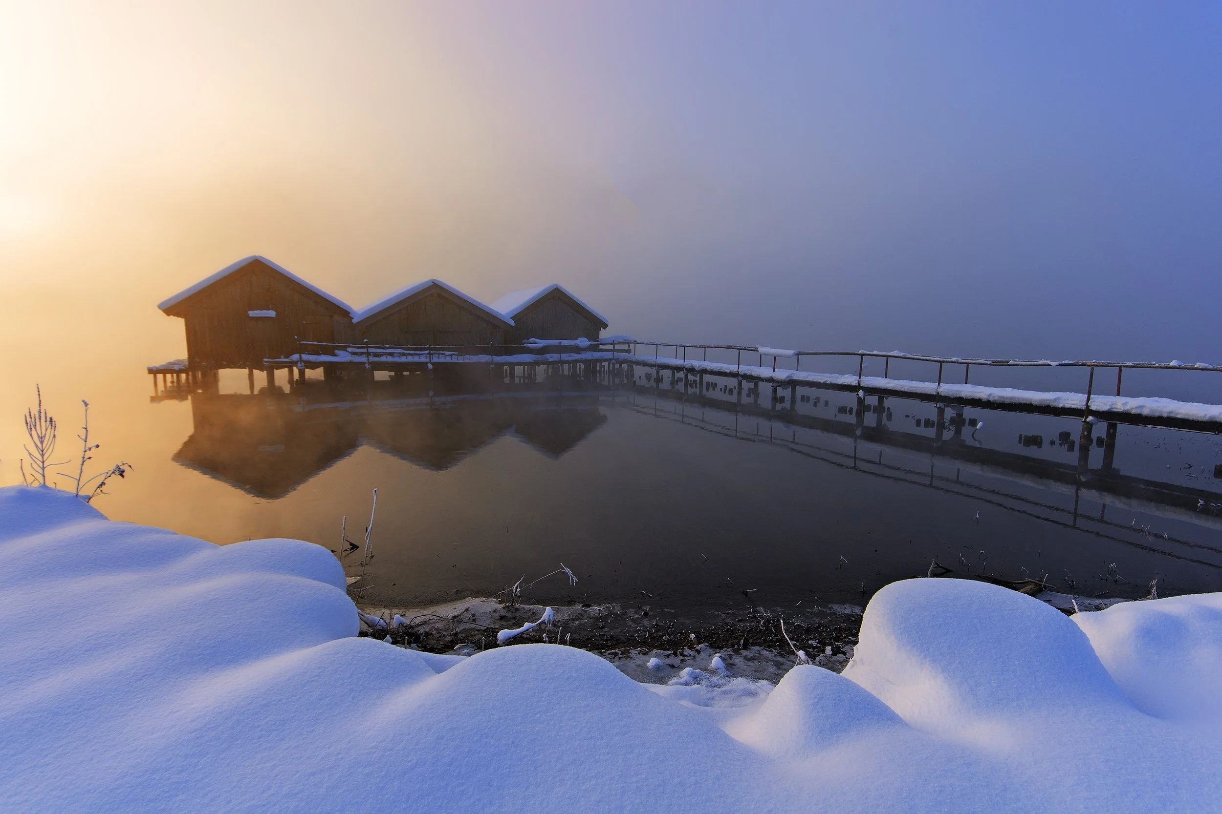 Three wooden houses on stilts over calm water with snow-covered ground in foreground, misty atmosphere, and sunrise or sunset glow in background. Taken in deep winter at Kochelsee, Bavaria.