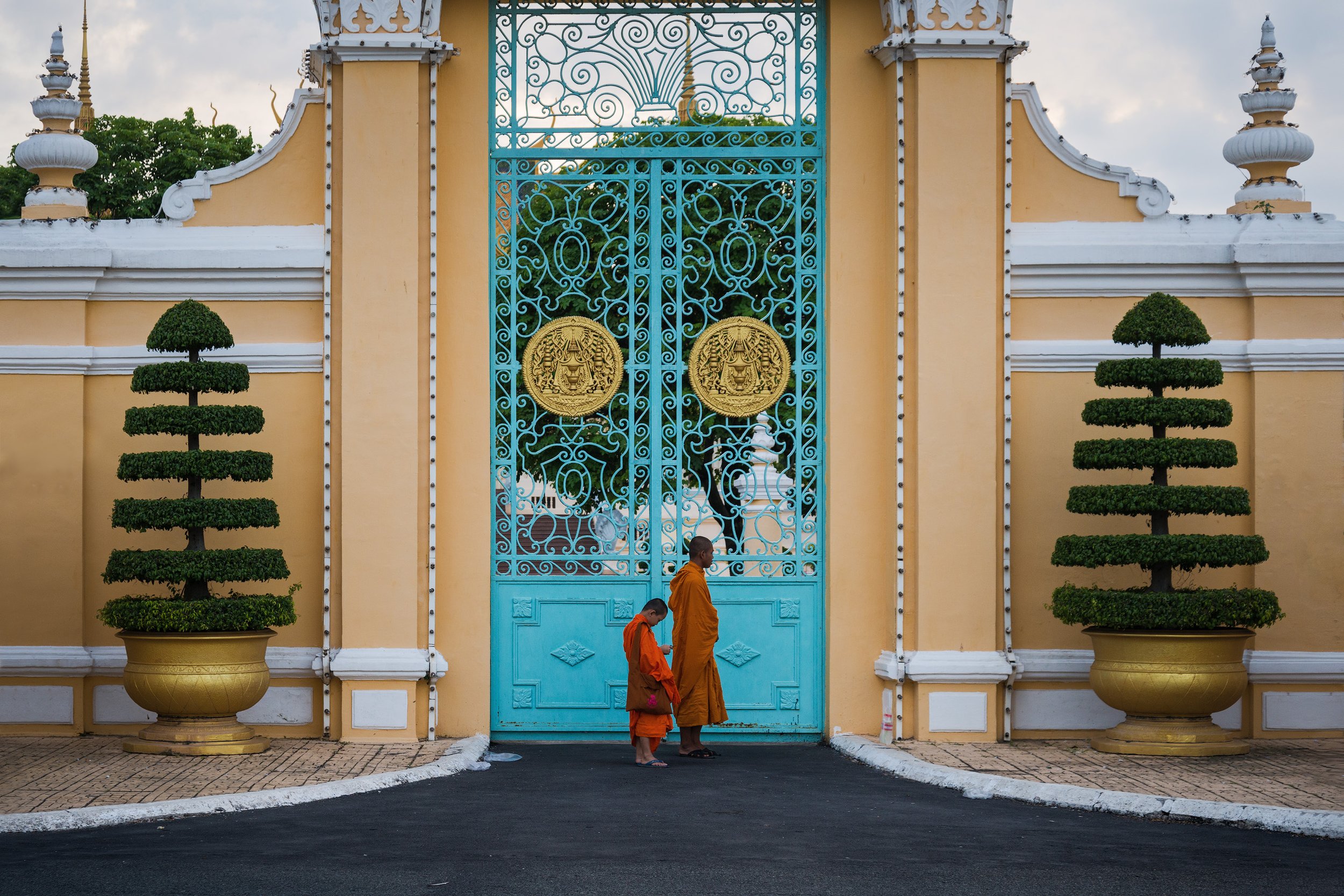 Two monks, one adult and one child, dressed in traditional orange robes, standing in front of a large ornate blue gate decorated with gold medallions. The gate is set into a yellow wall with white trim, flanked by symmetrical potted topiary trees.