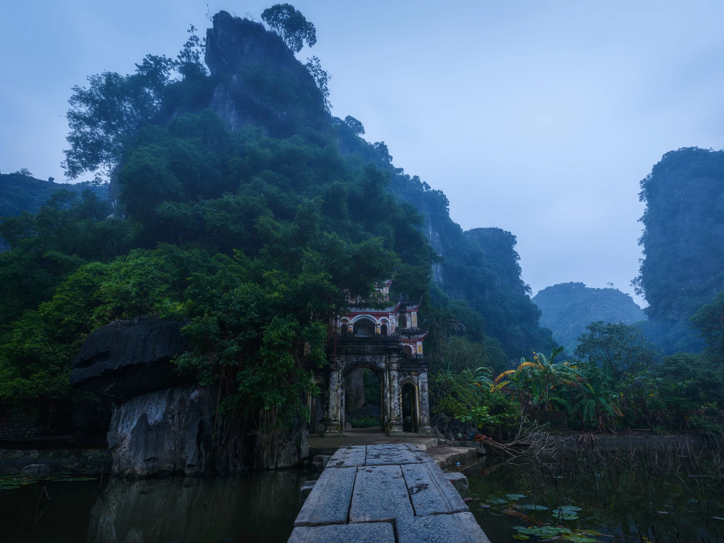Ancient stone archway on a narrow path leading to a traditional building surrounded by lush green trees, set against tall, forested mountains under a cloudy sky. Viet Nam, Vietnam.