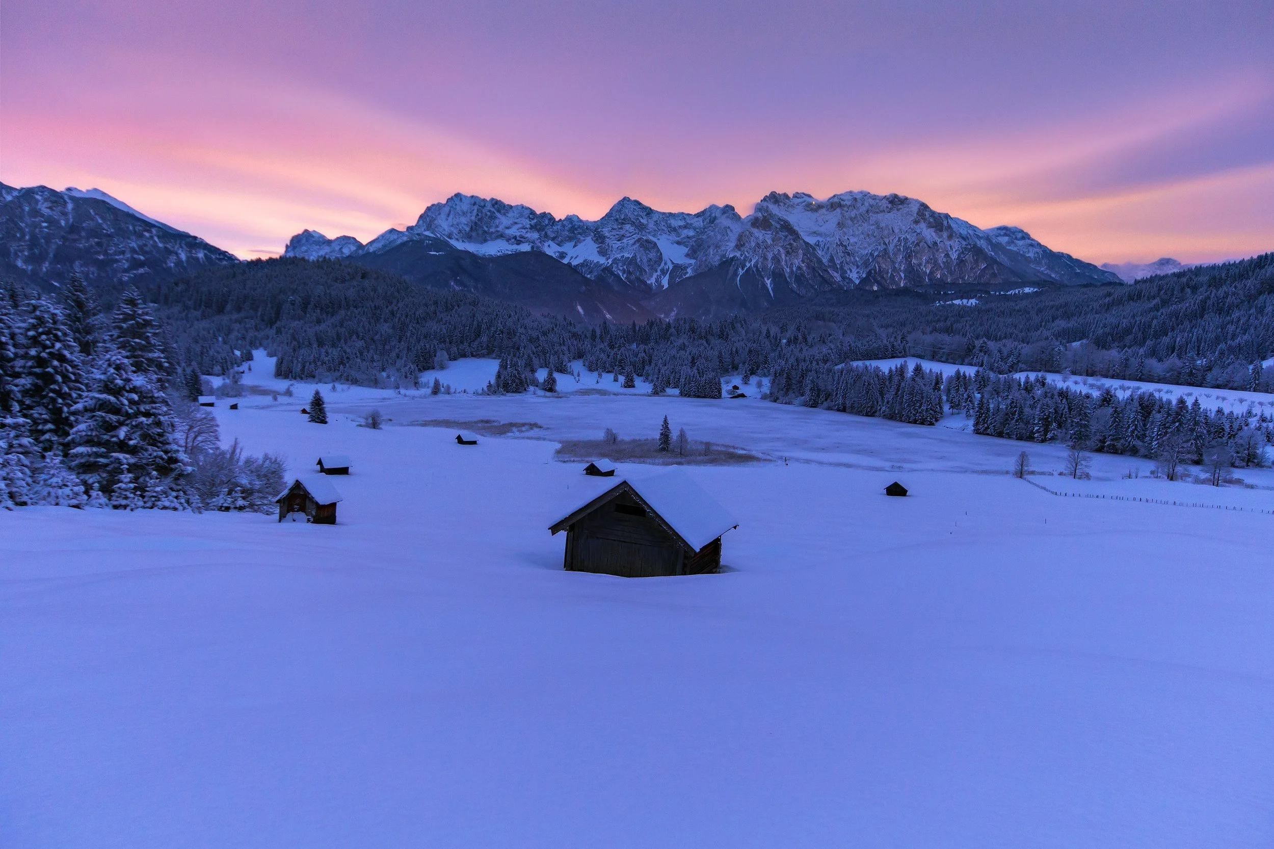 Snow-covered landscape with scattered wooden cabins, dense forest, and mountains in the background during sunset with pink and purple sky.