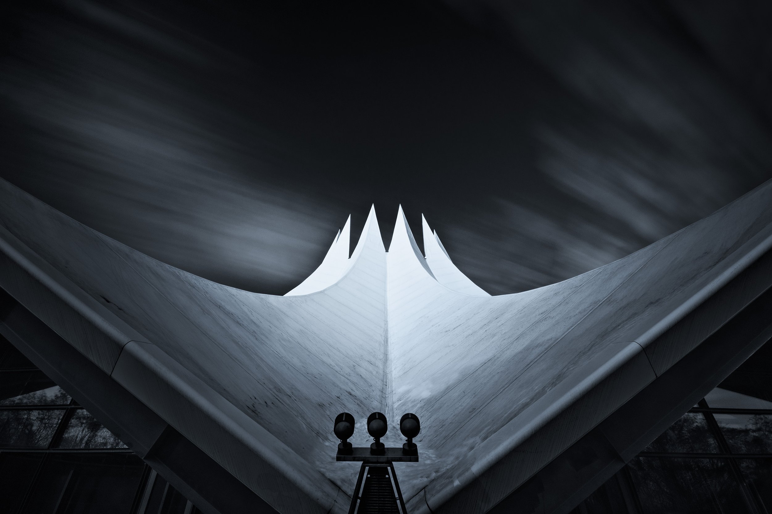 Low-angle view of a modern architectural structure with sharp, white peaks and a curved base, set against a dark, cloudy sky.