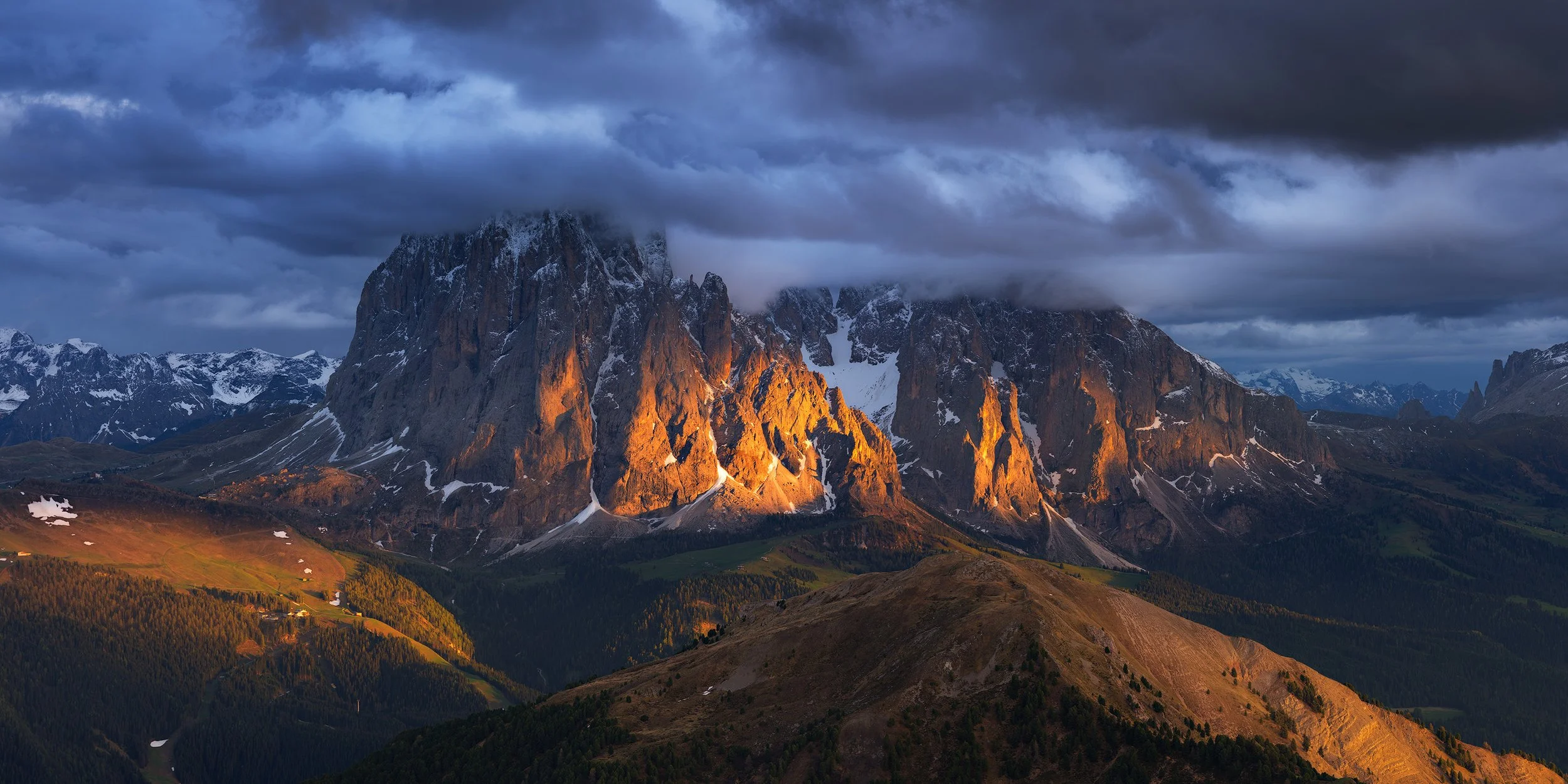 Mountain range under dark cloudy sky, with sunlight illuminating the rugged peaks and snow patches on the slopes.