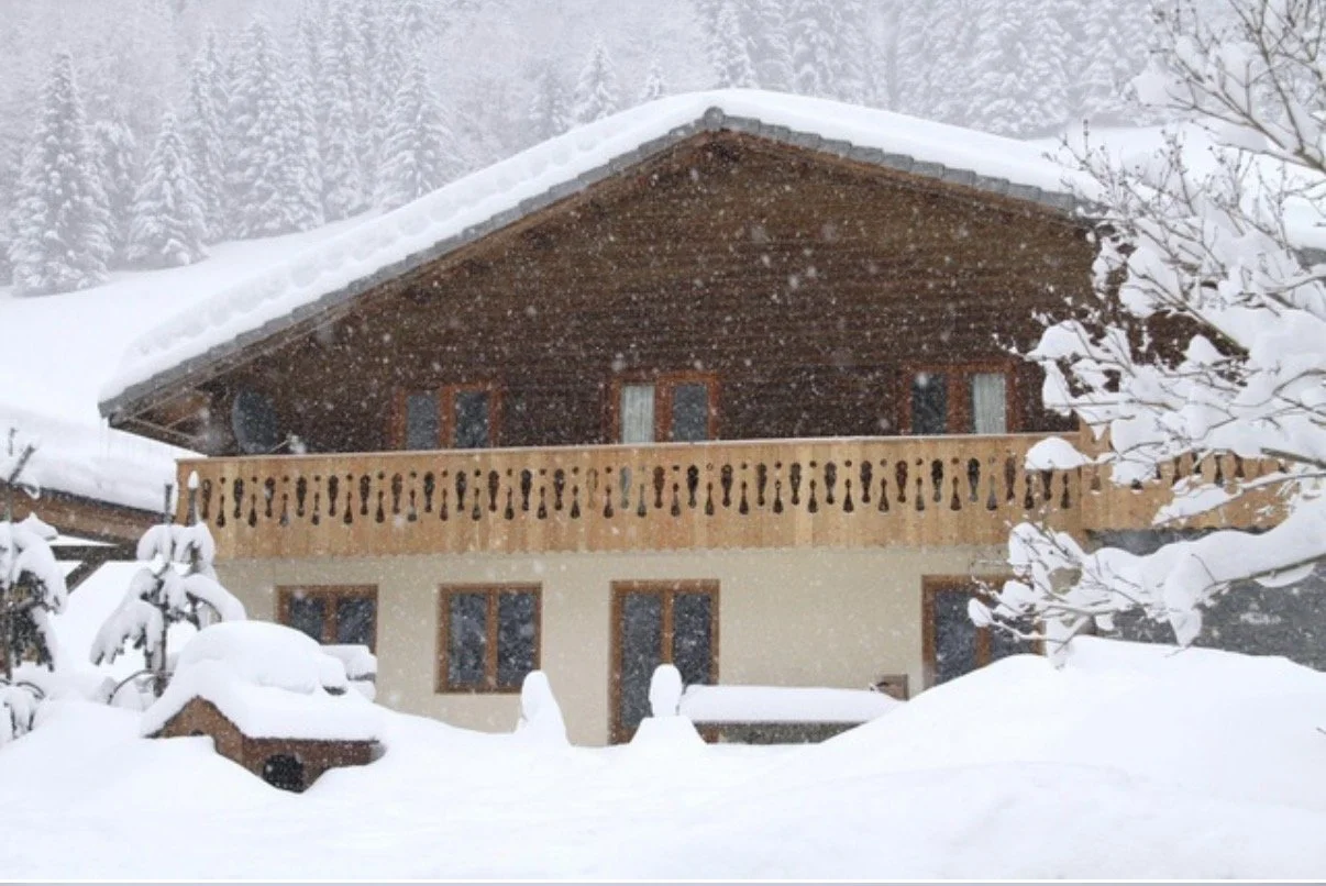 A snow-covered house with a wooden balcony and a sloped roof in a snowy landscape with trees in the background.