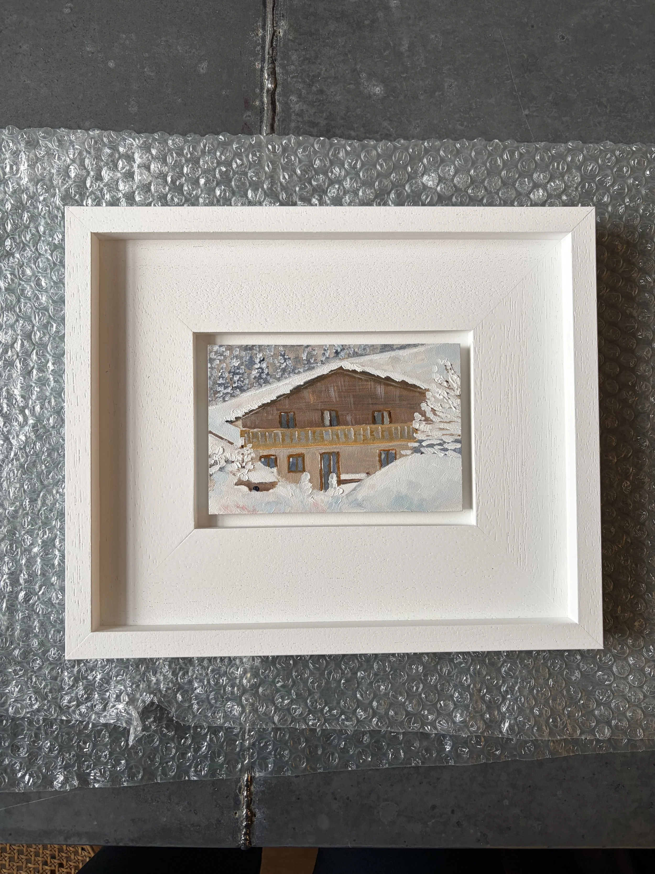 A framed painting of a snow-covered house with a brown roof and a wooden balcony in a winter landscape.