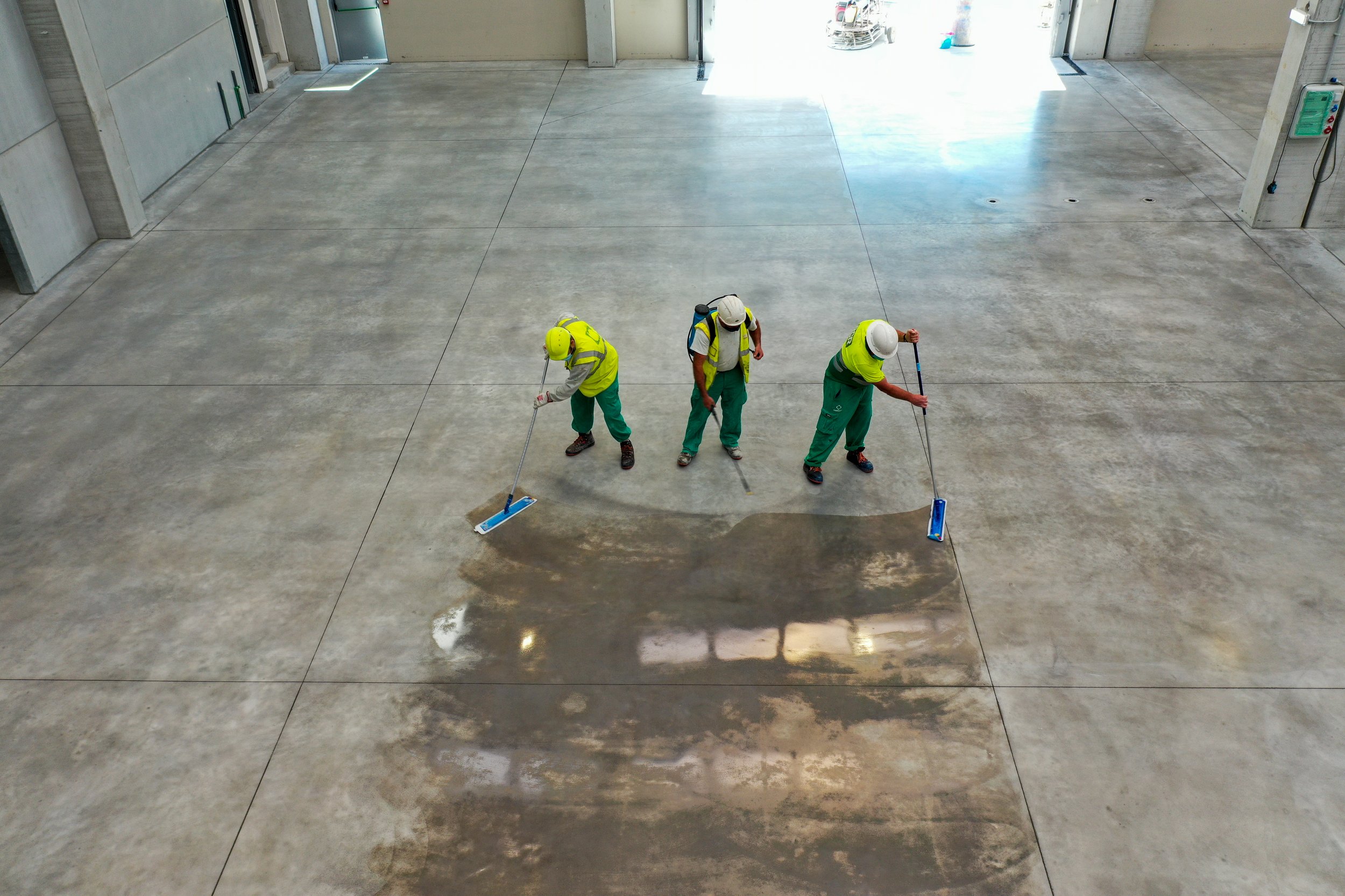 Three workers in yellow safety vests and white helmets are cleaning the polished concrete floor in a large indoor space using mops.