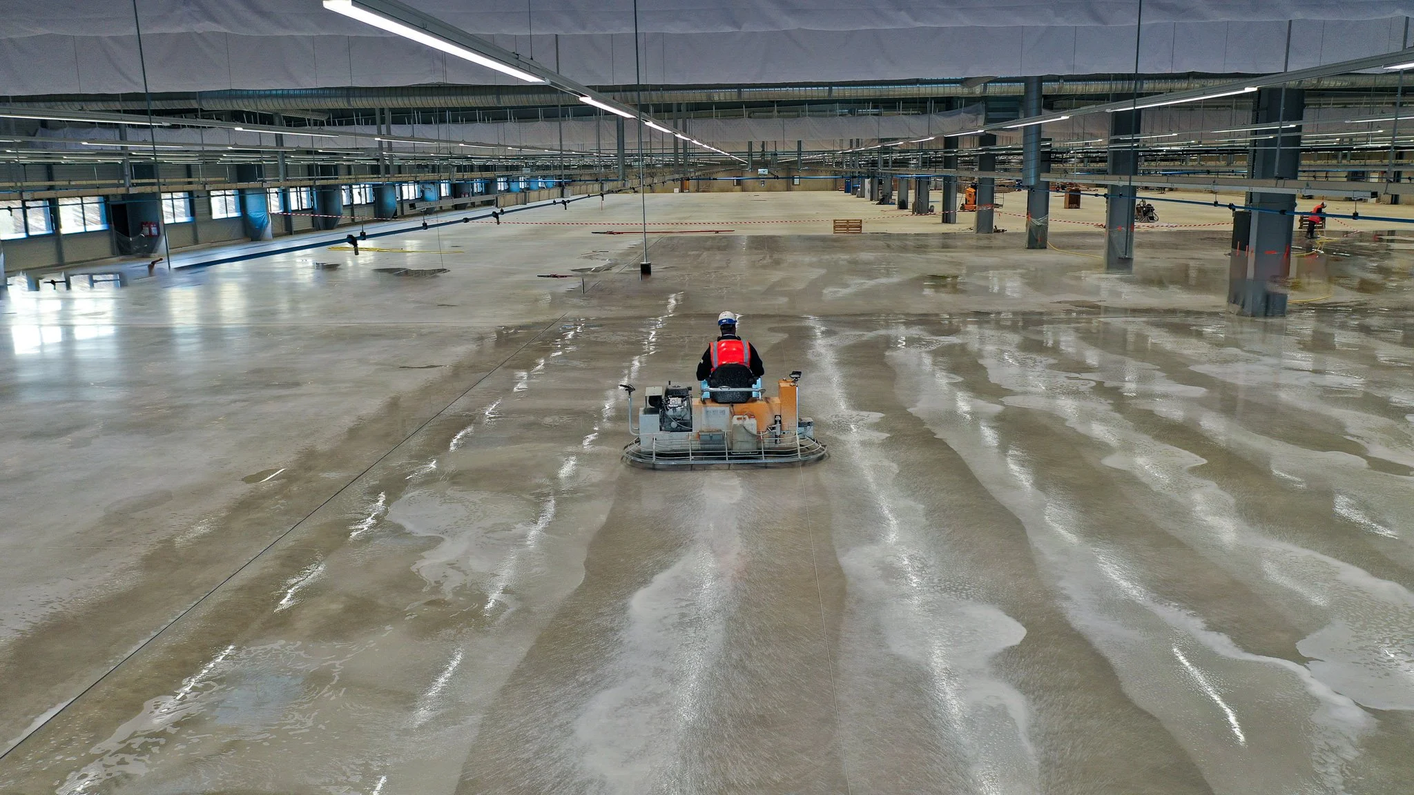 Worker operating a floor polishing machine in a large, empty indoor space with concrete floor and industrial ceiling.