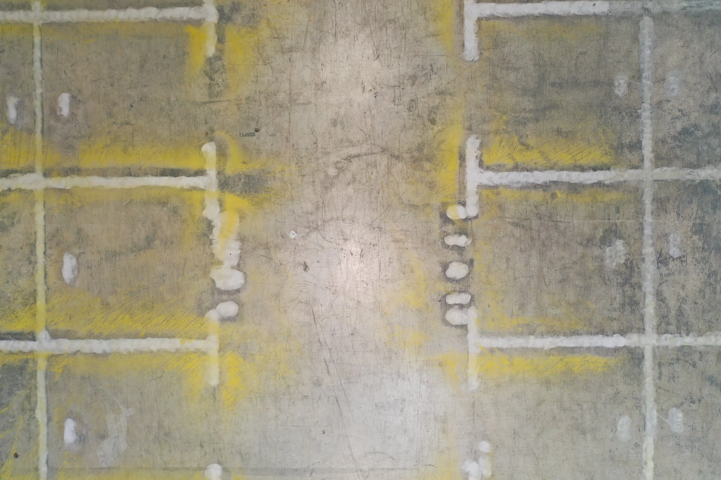 Close-up photograph of a concrete or stone floor with yellow and white markings, showing cement patches or glue spots, with the markings indicating a pattern or layout for tiles or bricks.
