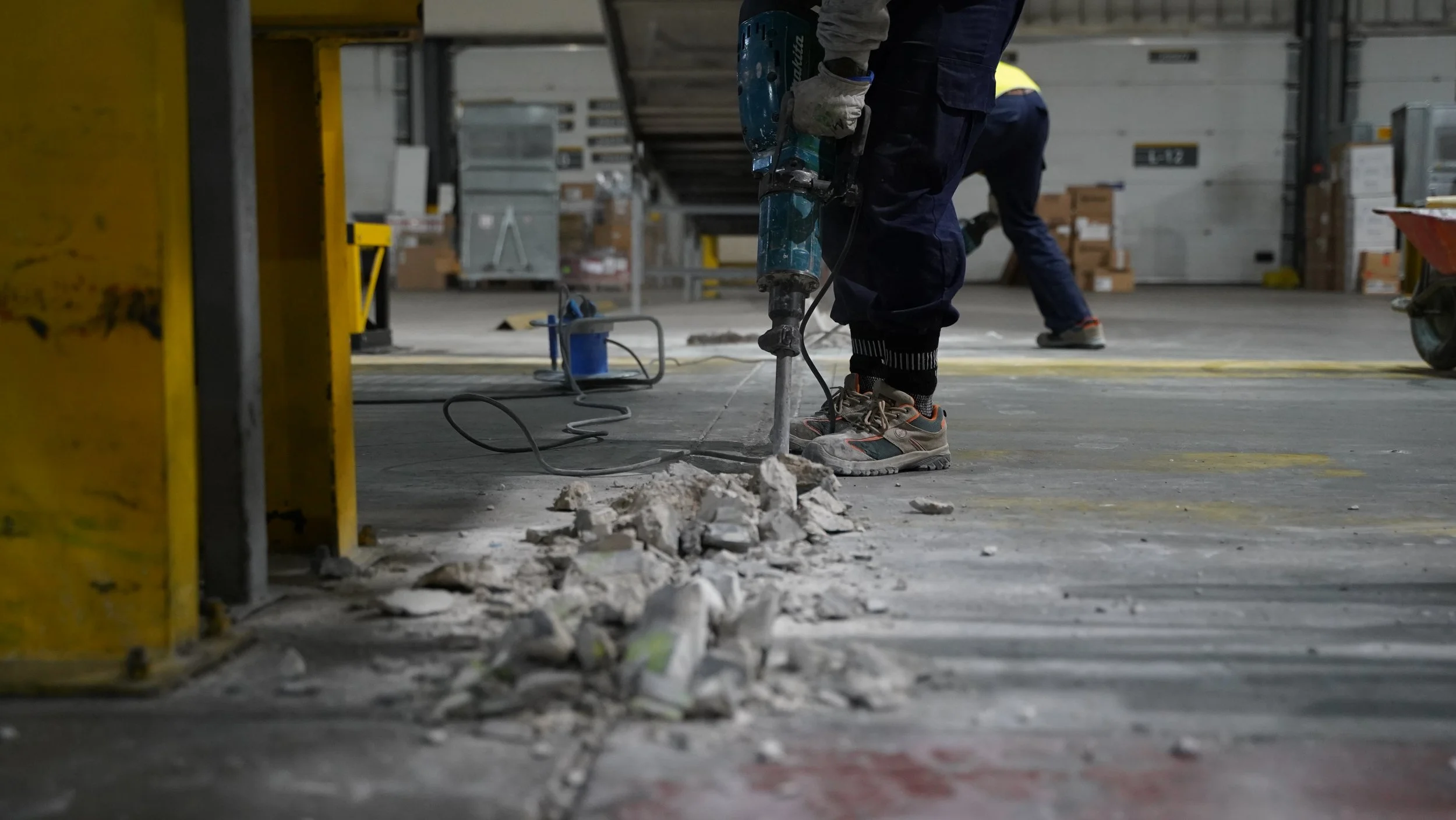 Worker using a jackhammer to break concrete on a warehouse floor with debris nearby, while another worker in the background walks past.