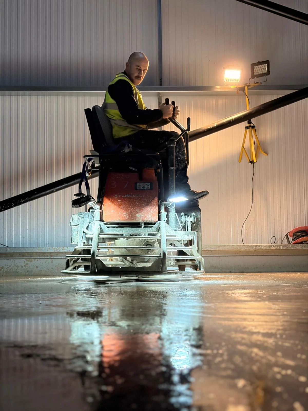 A worker operating a ride-on concrete floor grinder in an industrial setting, wearing a yellow safety vest, with equipment and lighting around him.