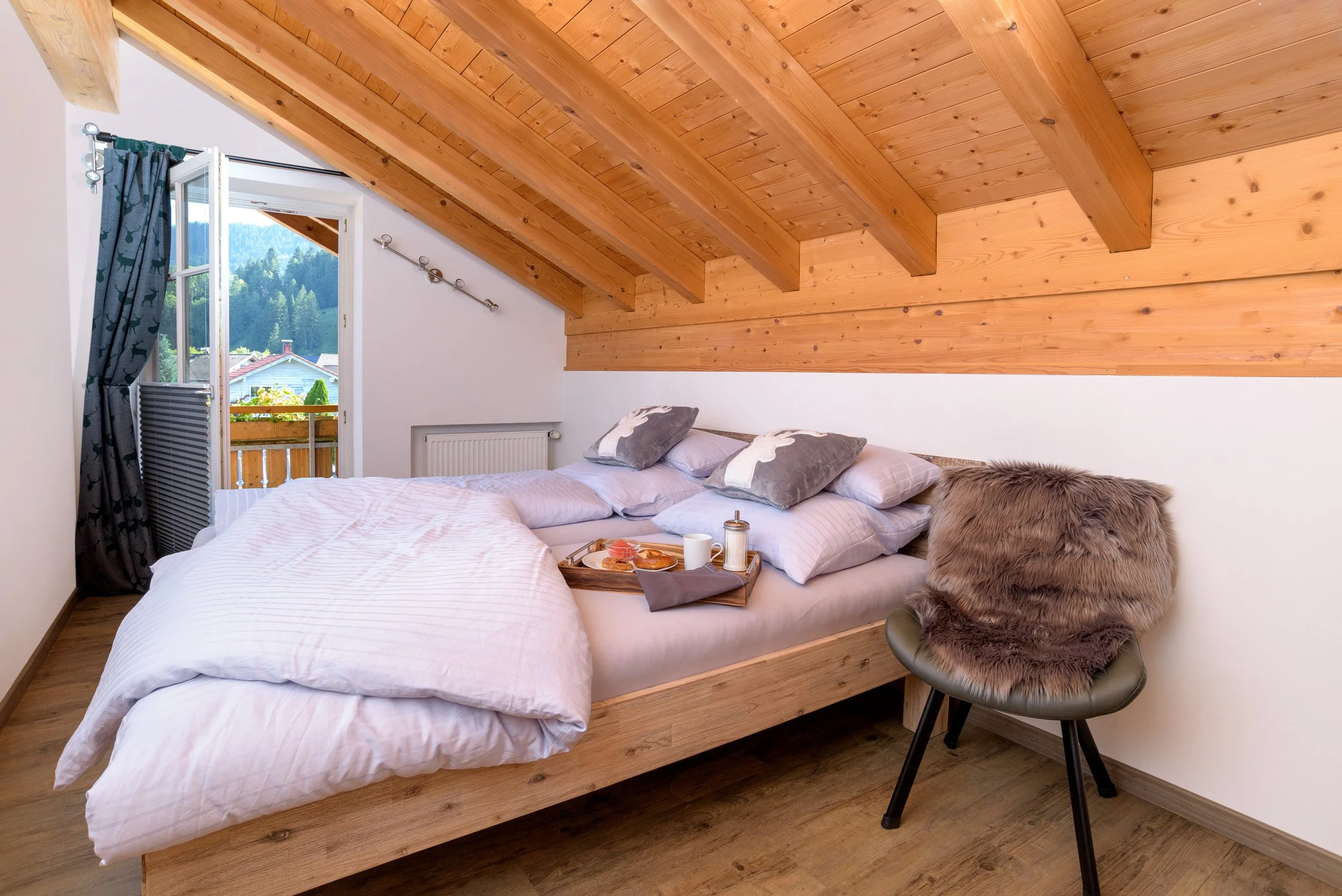 Berg Fux Ferienhaus Allgaeu Panorama Schlafzimmer mit einem Bett, das an eine Wand mit Holzdecke angrenzt. Das Bett hat weiße Bettwäsche und mehrere Kissen. Es gibt eine offene Balkontür mit Blick auf grüne Berge und eine kleine Terrasse.