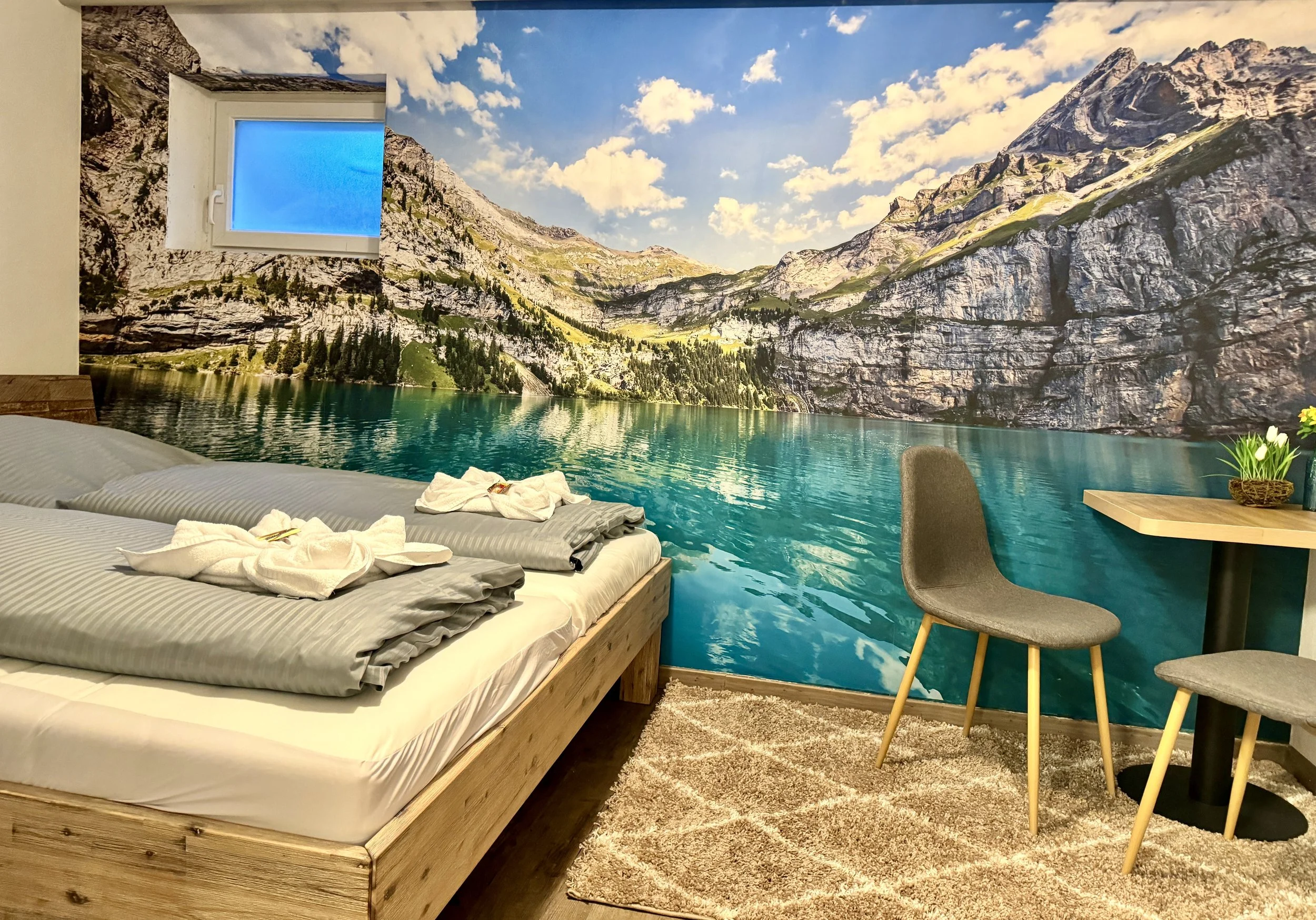 Berg Fux Ferienhaus Allgaeu Panorama Zimmer mit Bett, Frischwäsche, Wandbild einer Berglandschaft, kleiner Tisch mit Pflanze, Stuhl.