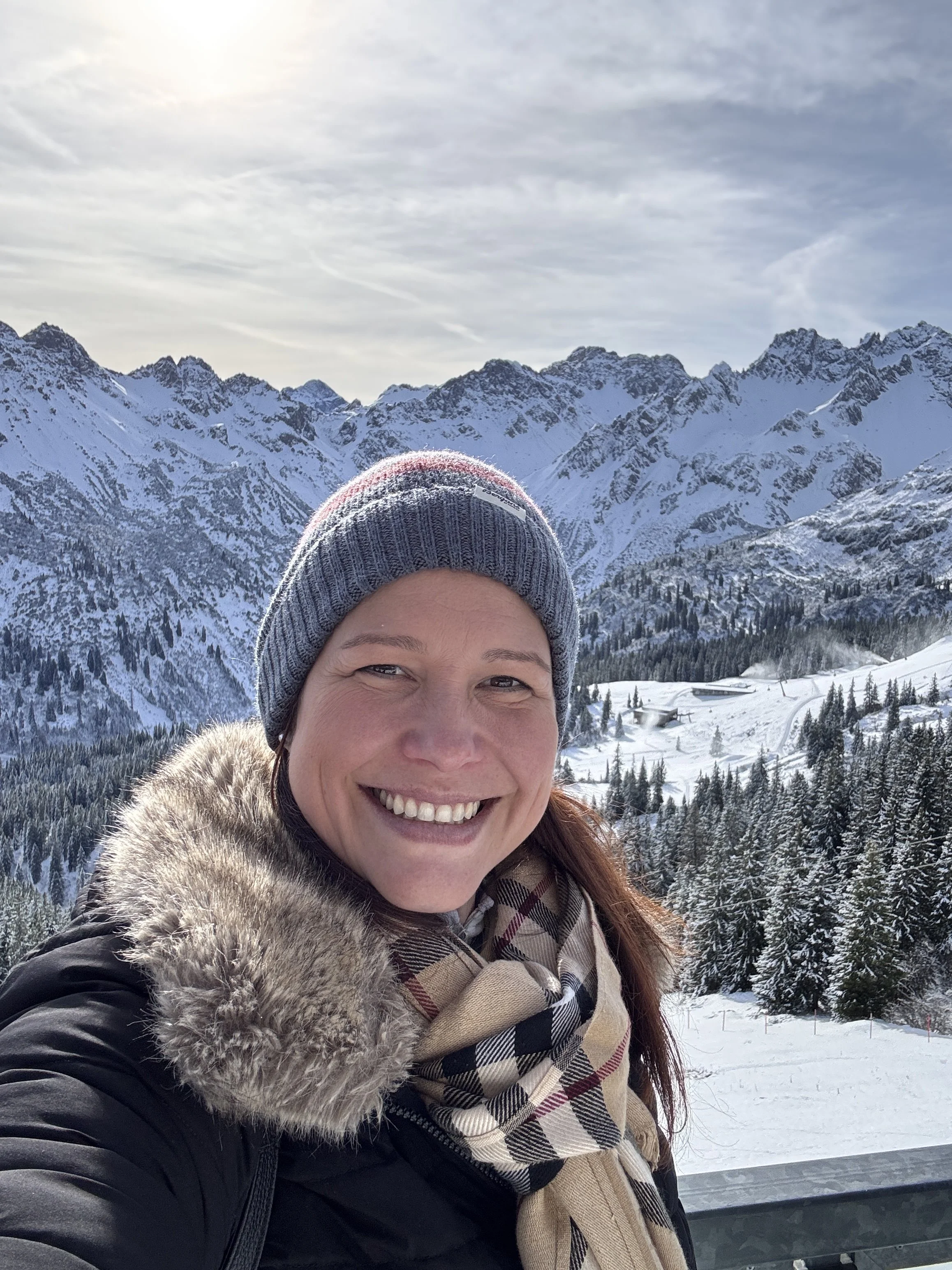 Berg Fux Eine lachende Frau mit braunen Haaren in Winterkleidung, inklusive Fellkapuzenjacke, Schal und Mütze, vor einer schneebedeckten Berglandschaft mit Wolken am Himmel.