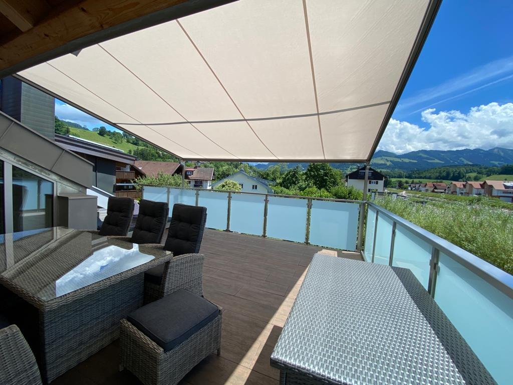 Berg Fux Ferienhaus mit Panorama Dachterasse mit Esstisch, Stühlen und Sonnenschutz mit Blick auf Berge und Häuser in einer ländlichen Umgebung.