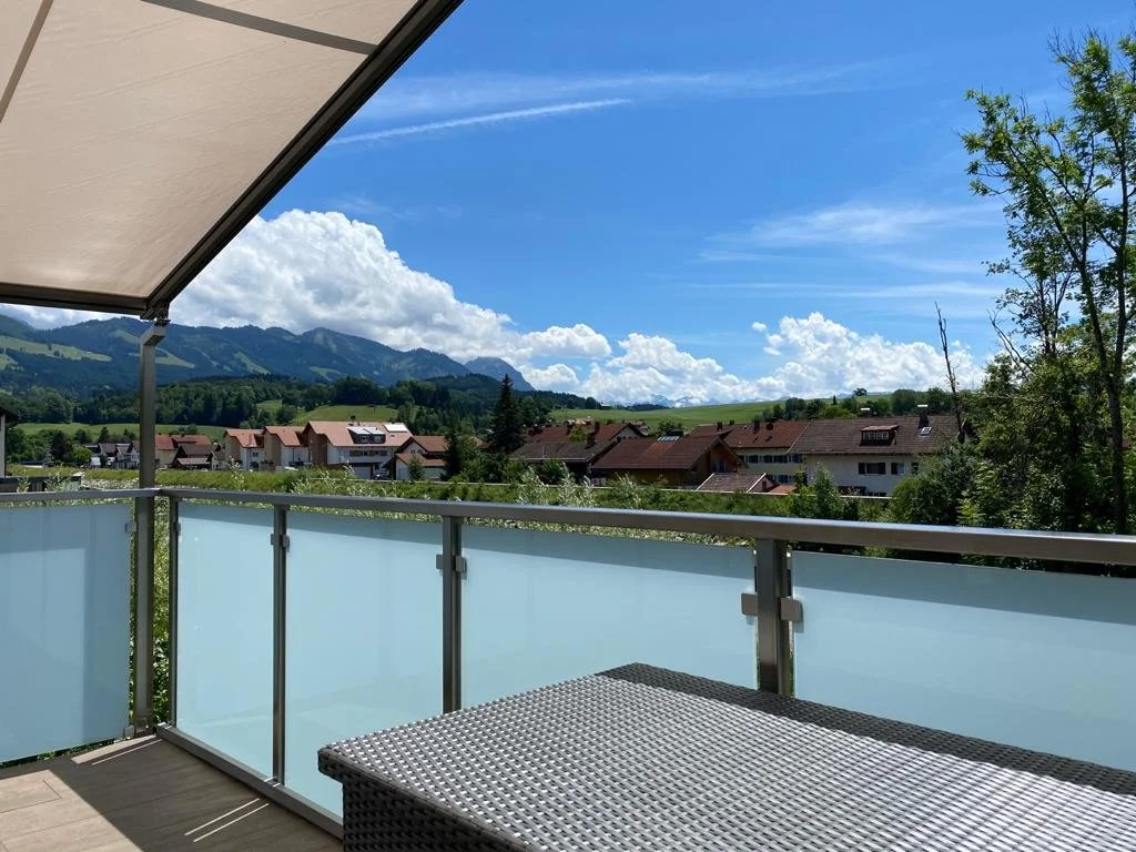 Berg Fux Ferienhaus Allgaeu Panorama Blick von einer Panorama Dachterasse auf eine ländliche Landschaft mit Häusern, grünen Hügeln und Bergen im Hintergrund, bei sonnigem Wetter.