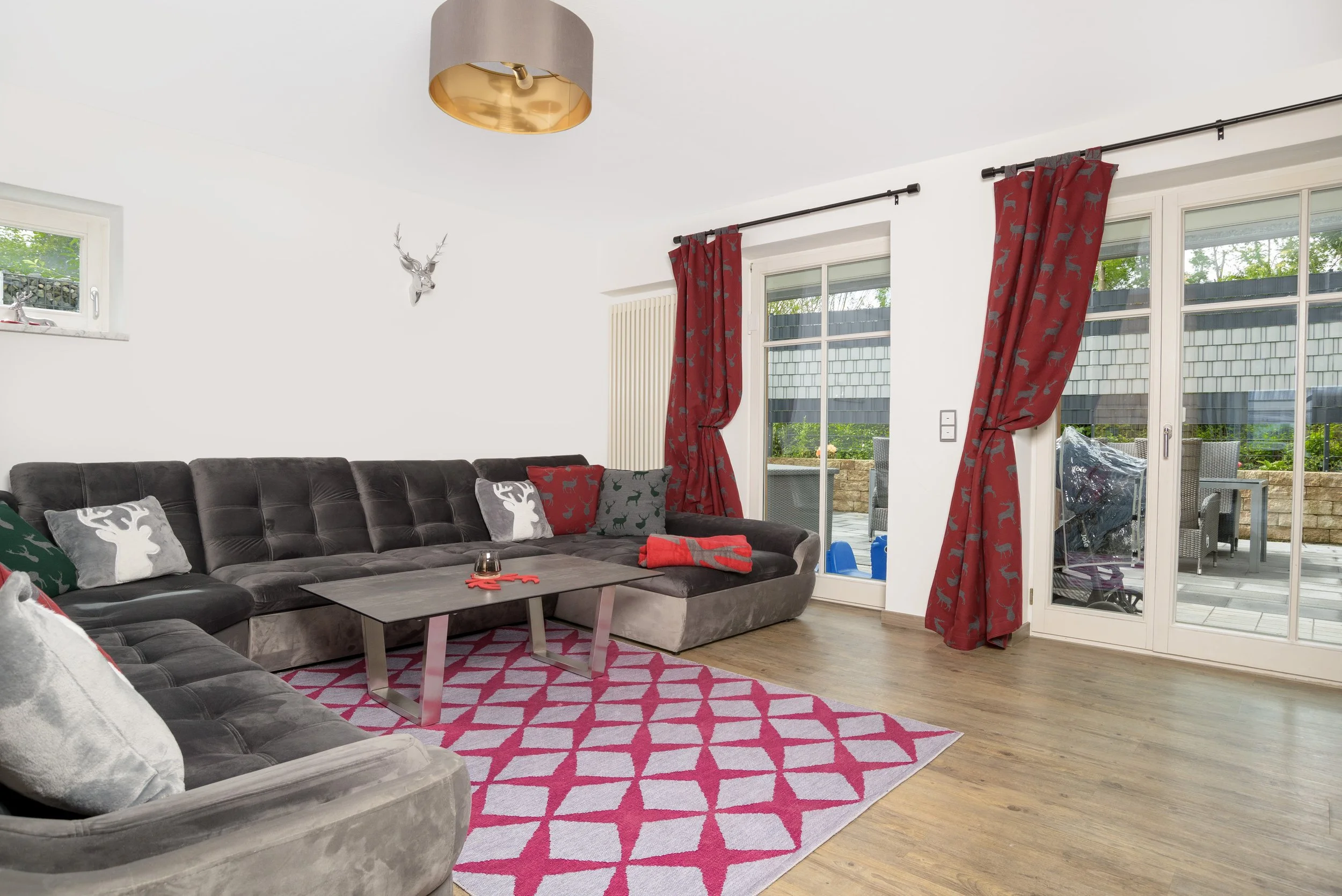 Berg Fux Ferienhaus Allgaeu Panorama Wohnzimmer mit grauer Couch, Weihnachtskissen, roter Teppich, roten Vorhängen mit Rentier-Muster, großen Fenstern mit Balkon-Terrasse, Laterne und einem Hirschkopf an der Wand.