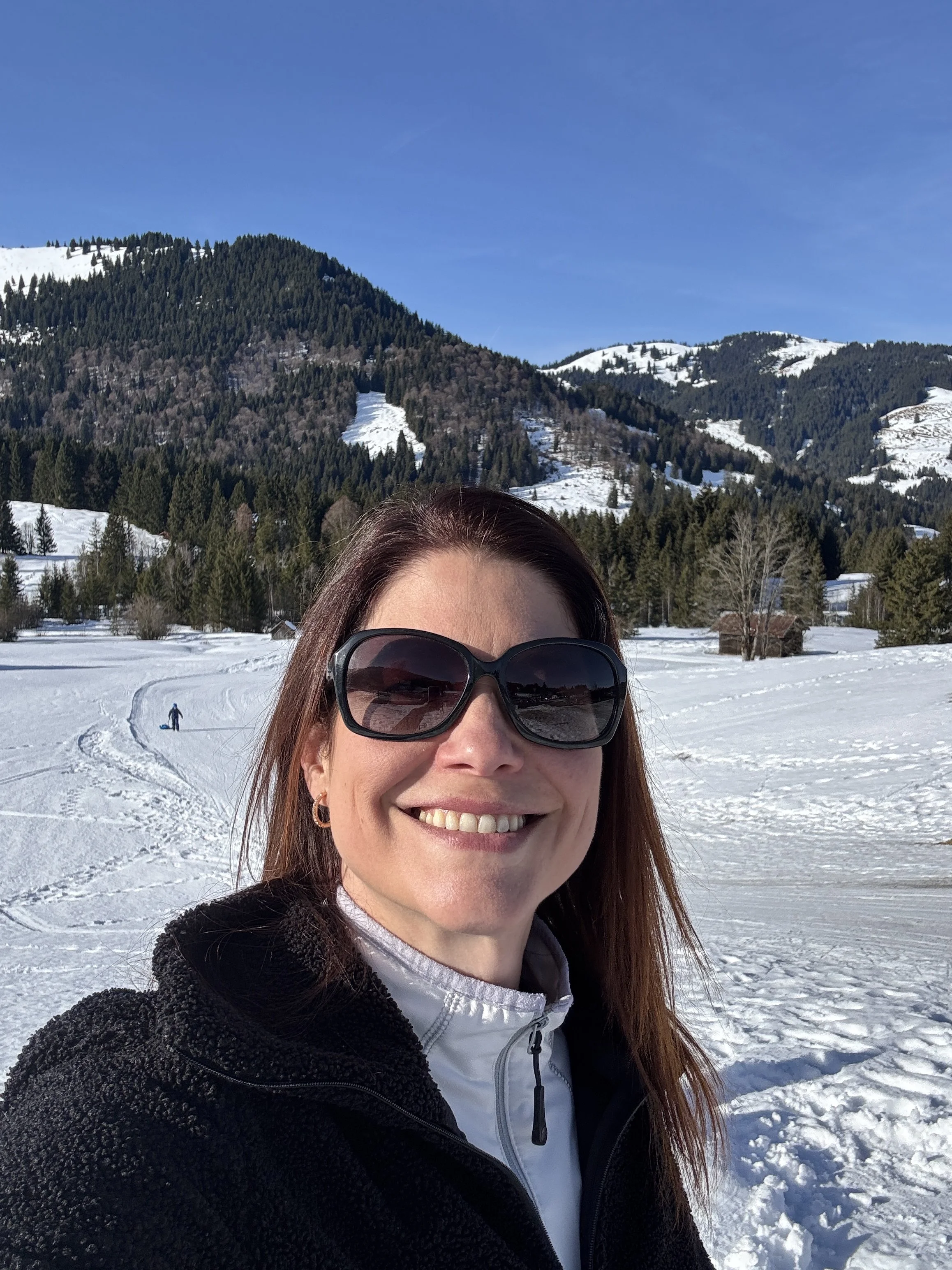 Berg Fux Stefanie Hilgers Inhaberin Eine lachende Frau mit dunklen Haaren und Sonnenbrille im Schnee vor einer winterlichen Berglandschaft mit Bäumen und Hügeln, blauer Himmel.