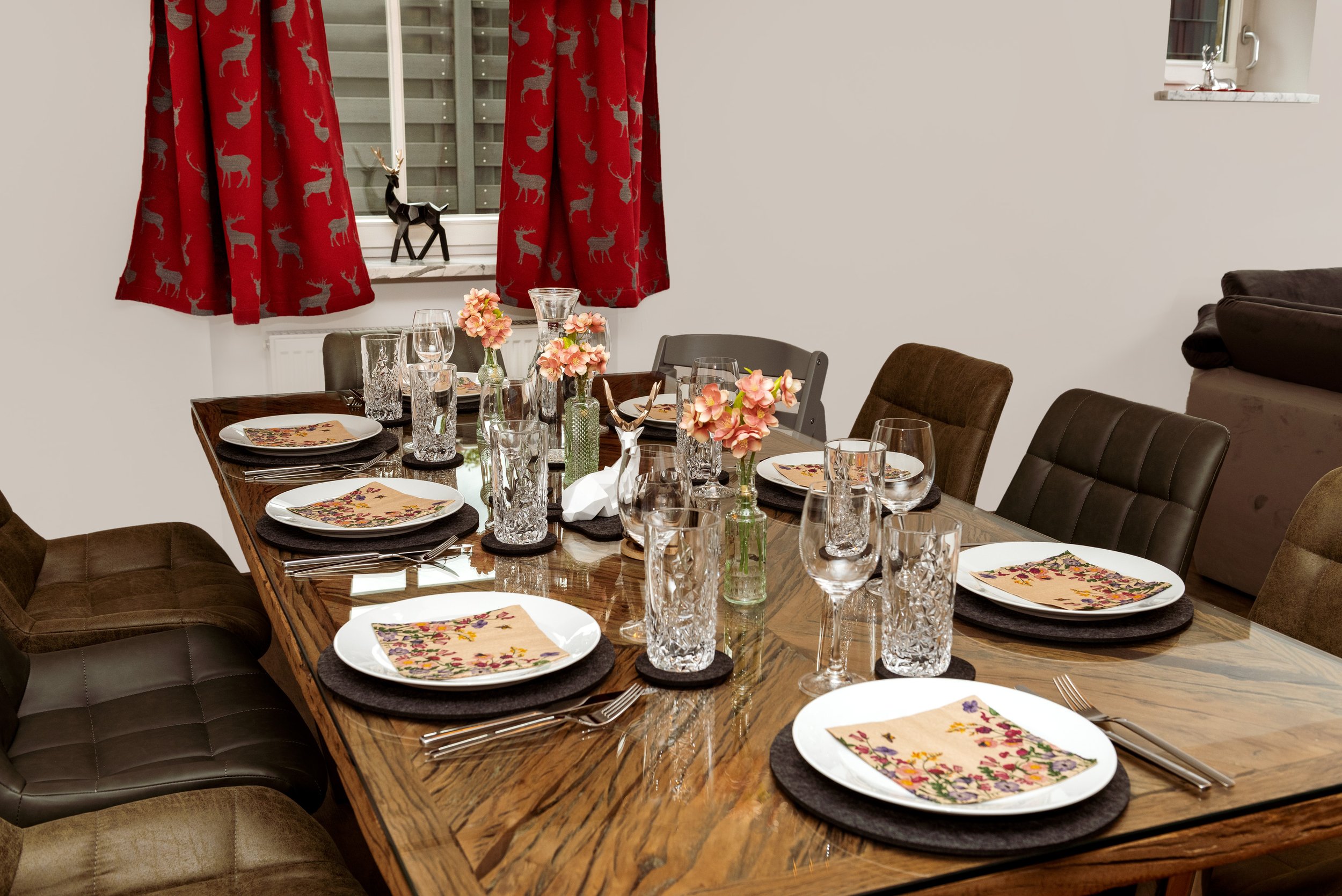 Berg Fux Ferienhaus Allgaeu Panorama Ein festlich gedeckter Esstisch mit weißen Tellern, bunten Servietten, Kristallgläsern und Blumen in Vasen, in einem Wohnzimmer mit roten Weihnachtsvorhängen mit Hirschmuster, Fenster und Sofas.