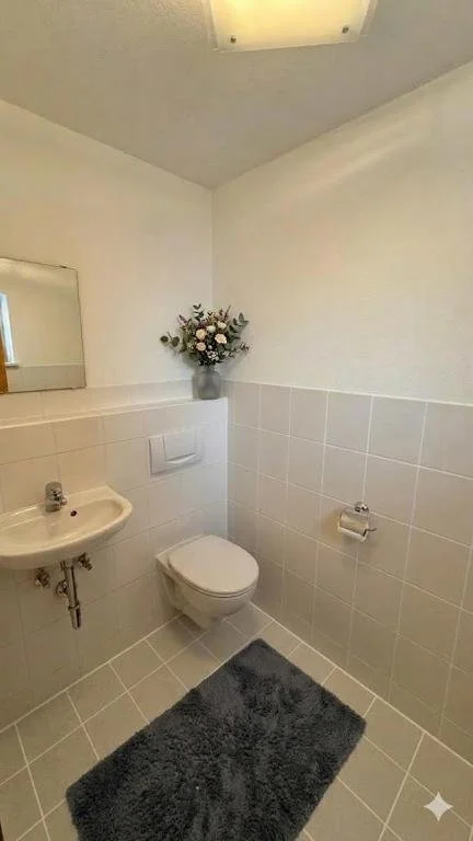 Kleines Badezimmer mit weißen Fliesen, einer Toilette, einem Waschbecken, einem Spiegel, einer Blumenstrauß auf der Ablage, einem grauen Badezimmerteppich und einer Deckenleuchte.