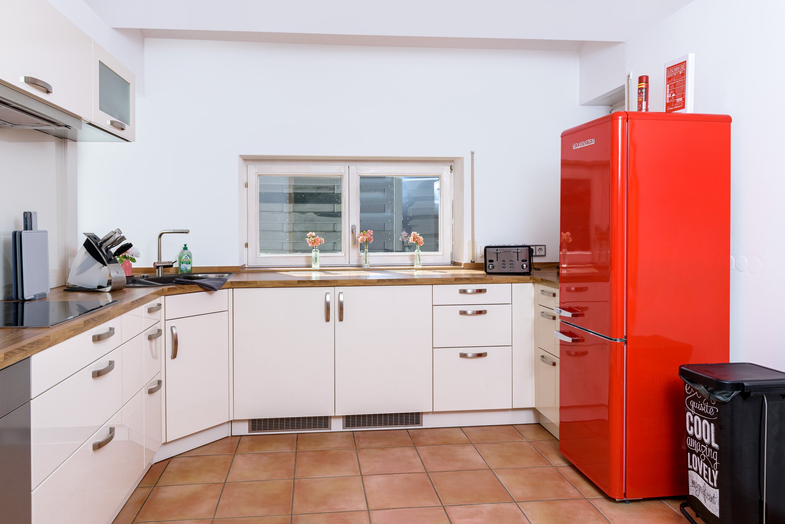 Berg Fux Ferienhaus Allgaeu Panorama Küche mit weißen Schränken, Holzarbeitsplatten, einem Fenster mit Blick nach draußen, einer roten Kühlschrank, kleinen Vasen mit Blumen auf der Fensterbank, einem Toaster, einem Geschirrhandtuch