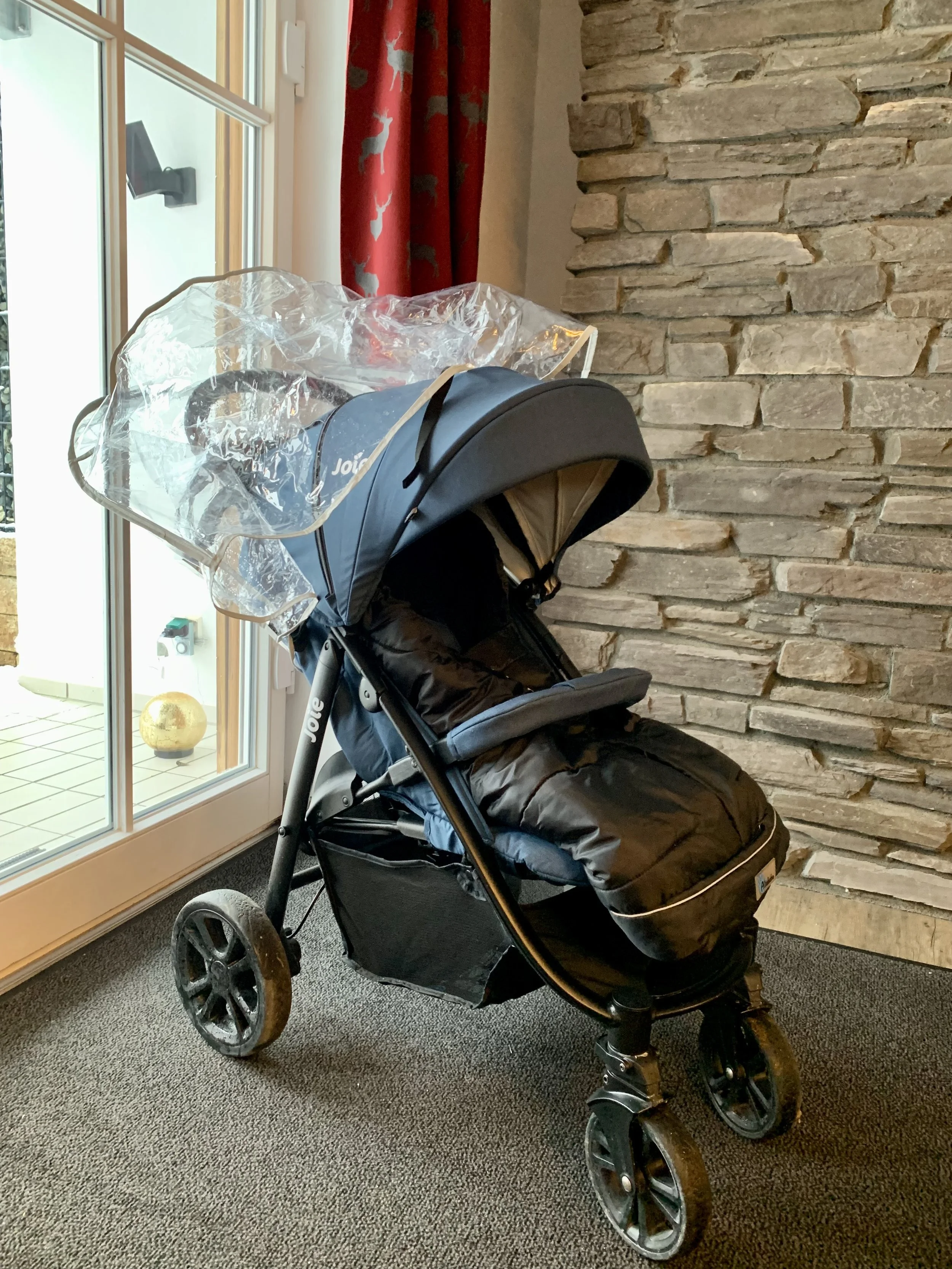 Kinderwagen mit Regenhaube vor einer Steinmauer in einem Innenraum, neben einem Glasfenster.