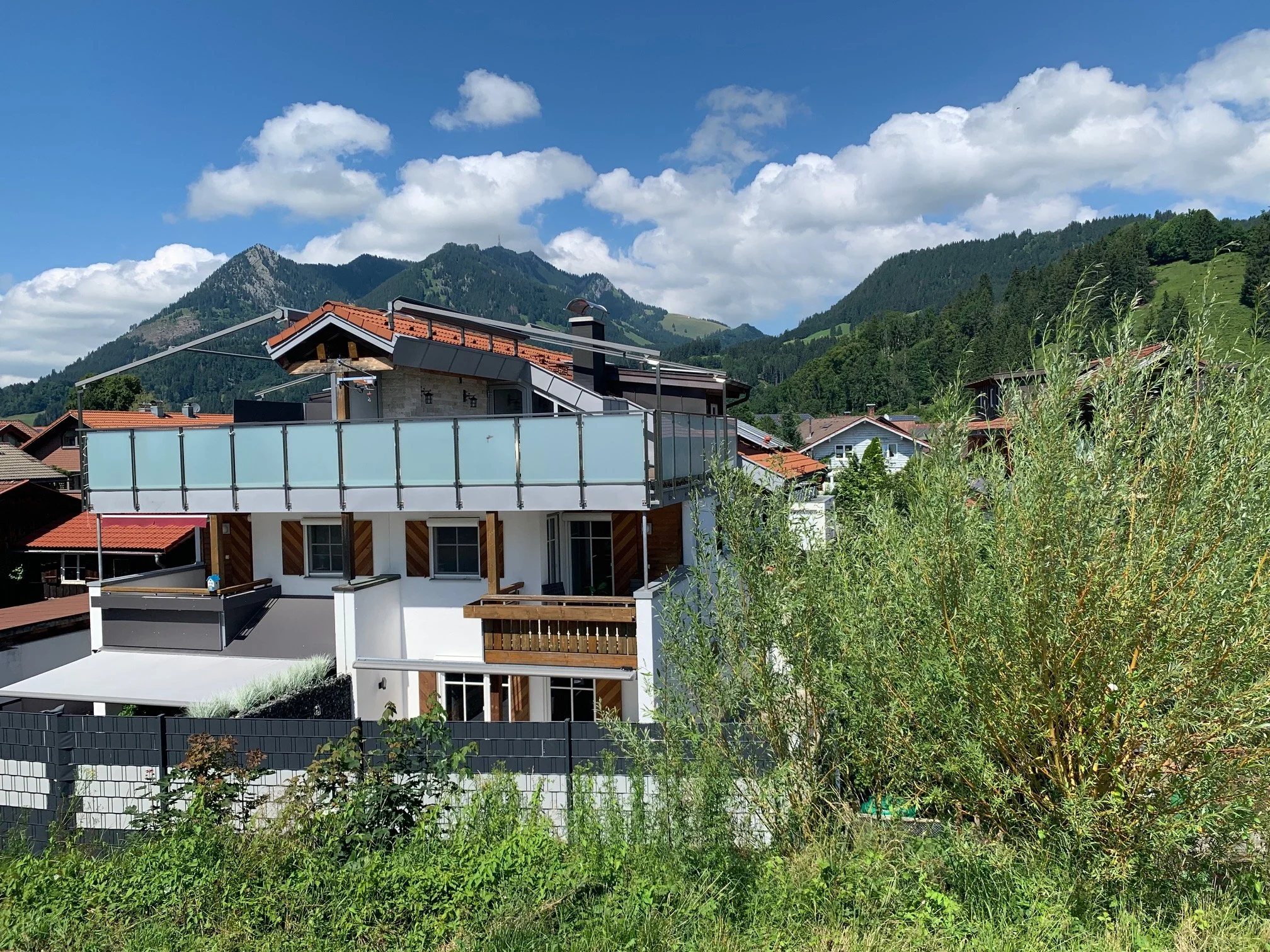 Berg Fux Ferienhaus Moderne Häuser mit Terrassen und Glasgeländern vor grünen Hügeln und Bergen unter einem blauen Himmel mit Wolken.