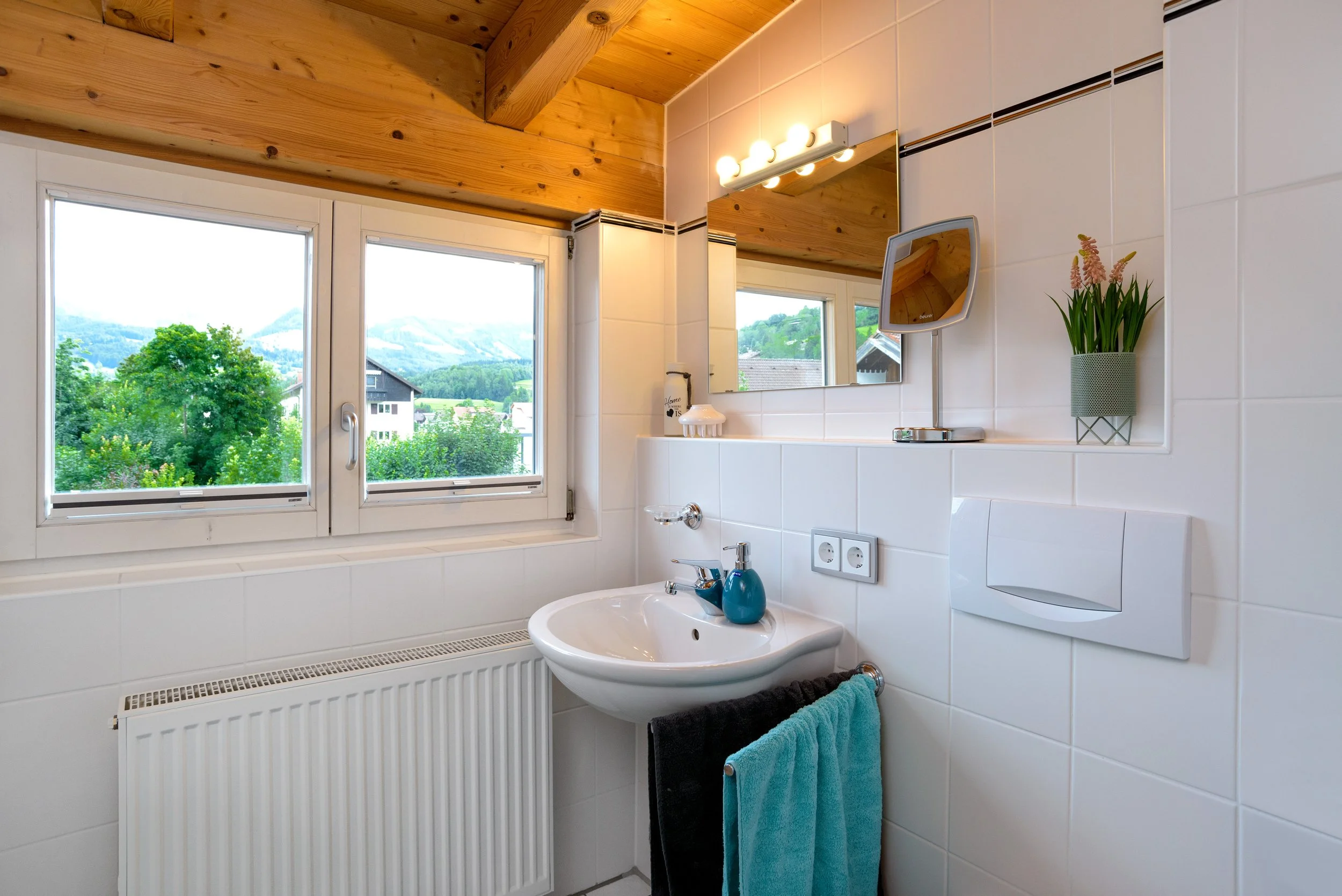 Berg Fux Ferienhaus Allgaeu Panorama Ein Badezimmer mit weißen Wandfliesen, einem Waschbecken mit einer blauen Seifenschale und Handtüchern, eine große Fensterfront mit Blick auf grüne Bäume und Berge im Hintergrund sowie eine Holzdecke.