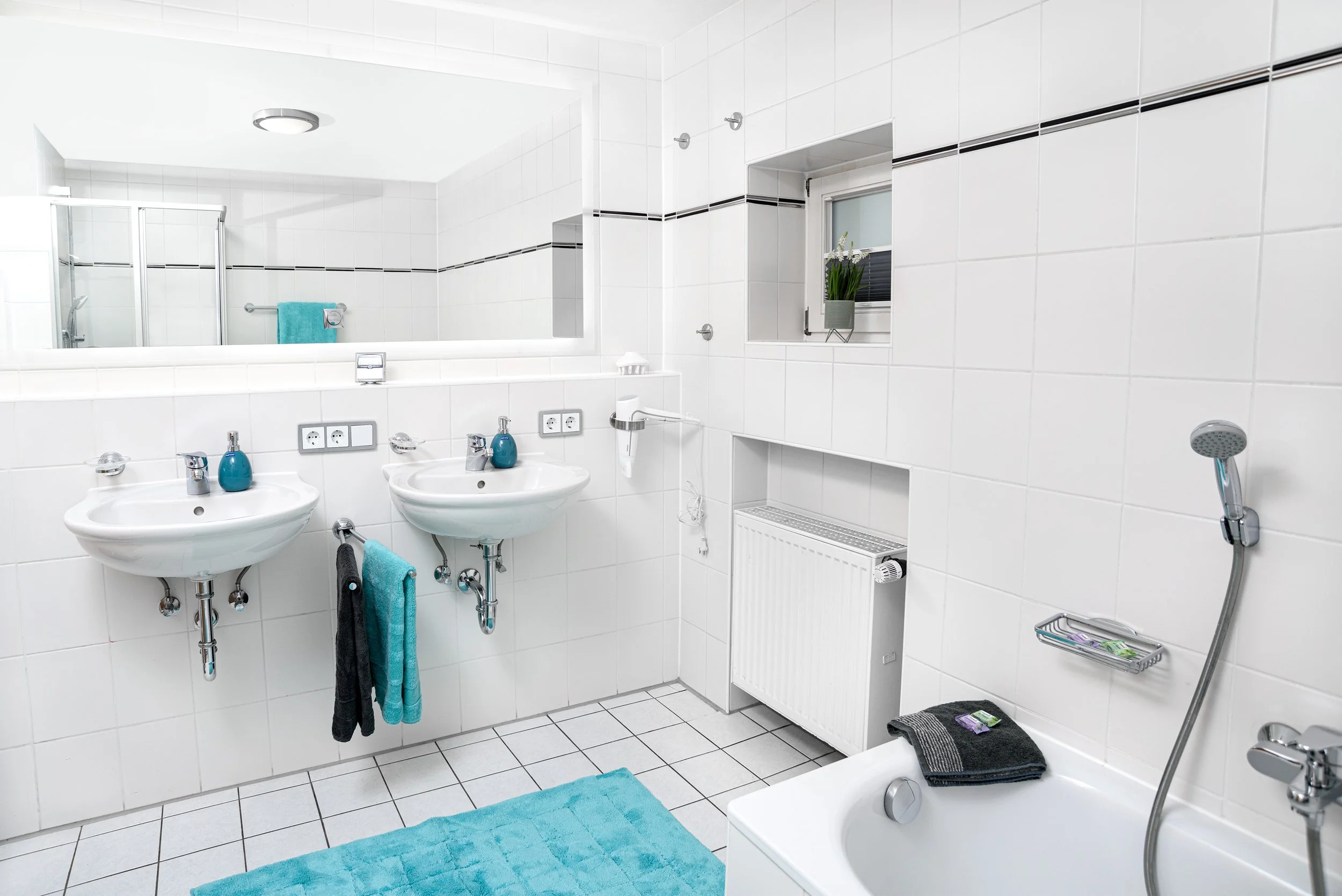 Berg Fux Ferienhaus Allgaeu Panorama Ein weißes Badezimmer mit zwei Waschbecken, einer Badewanne und einer Dusche, weißen Fliesen und blauen Handtüchern.