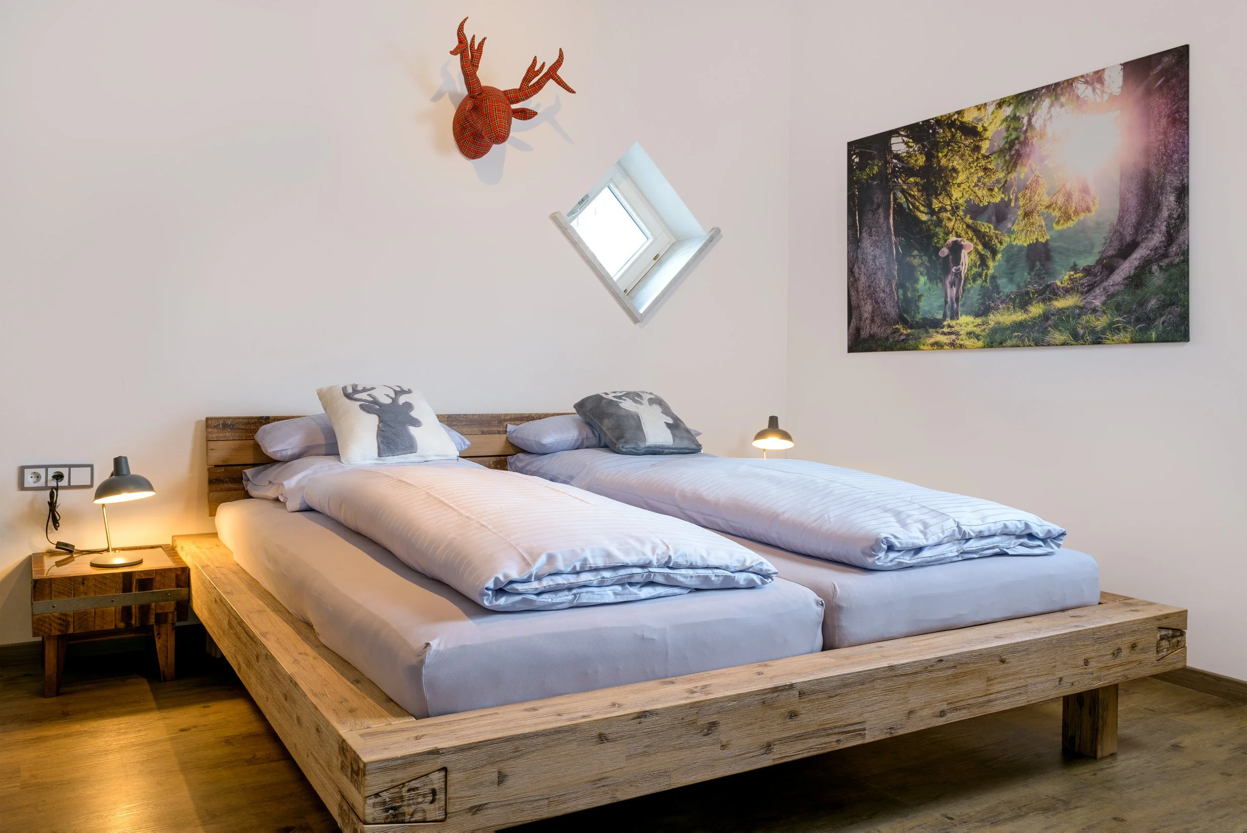 iBerg Fux Ferienhaus Allgaeu Panorama Ein Schlafzimmer mit zwei Einzelbetten aus Holz, jeweils mit weißen Bettwäsche und Kissen mit Hirschmuster. Über dem Bett befindet sich einen Wanddekoration in Form eines roten Hirschkopfes mit Geweih.