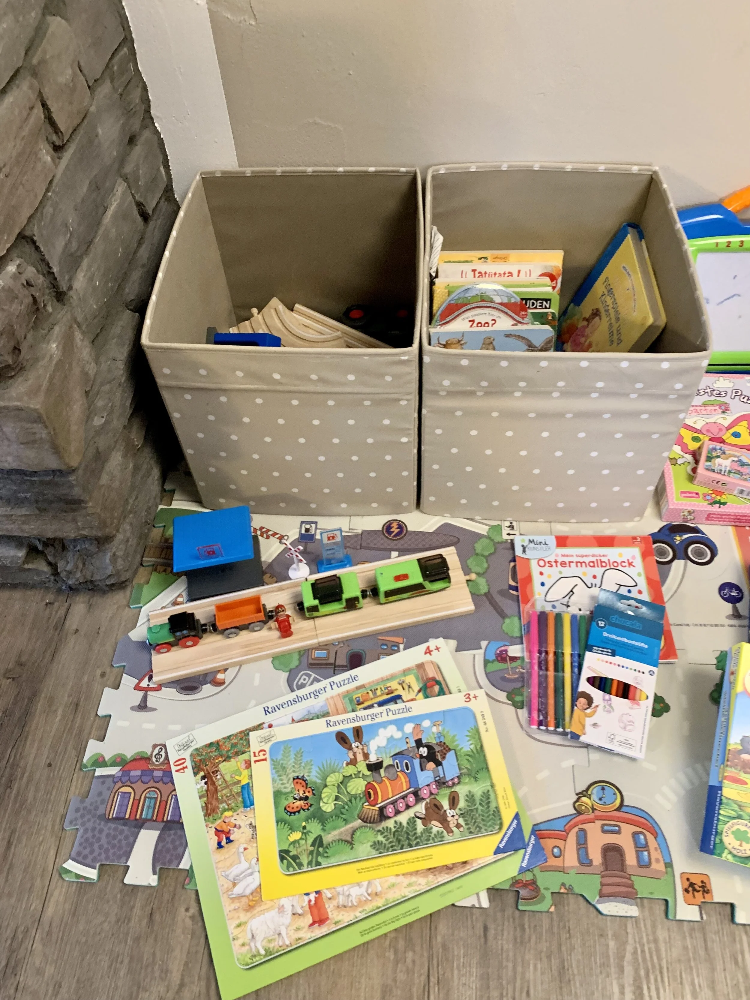 Berg Fux  Sammlung von Kinderpuzzles, Bücher und Bastelmaterial auf einem Boden. Im Hintergrund eine graue Wand und eine Steinmauer.