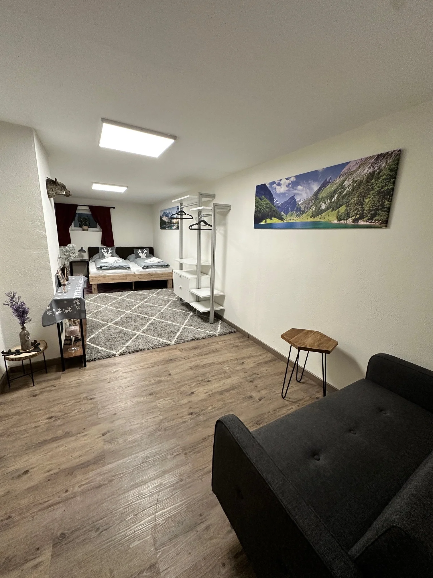 Berg Fux Ferienhaus Allgaeu Panorama  Wohnzimmer und Schlafzimmer mit Holzfußboden, grauem Sofa, kleinen Beistelltischen, einer Wanddekoration mit Berglandschaft, einem Bett mit weißen Bettwäsche und Kissen, einem Kleiderständer,